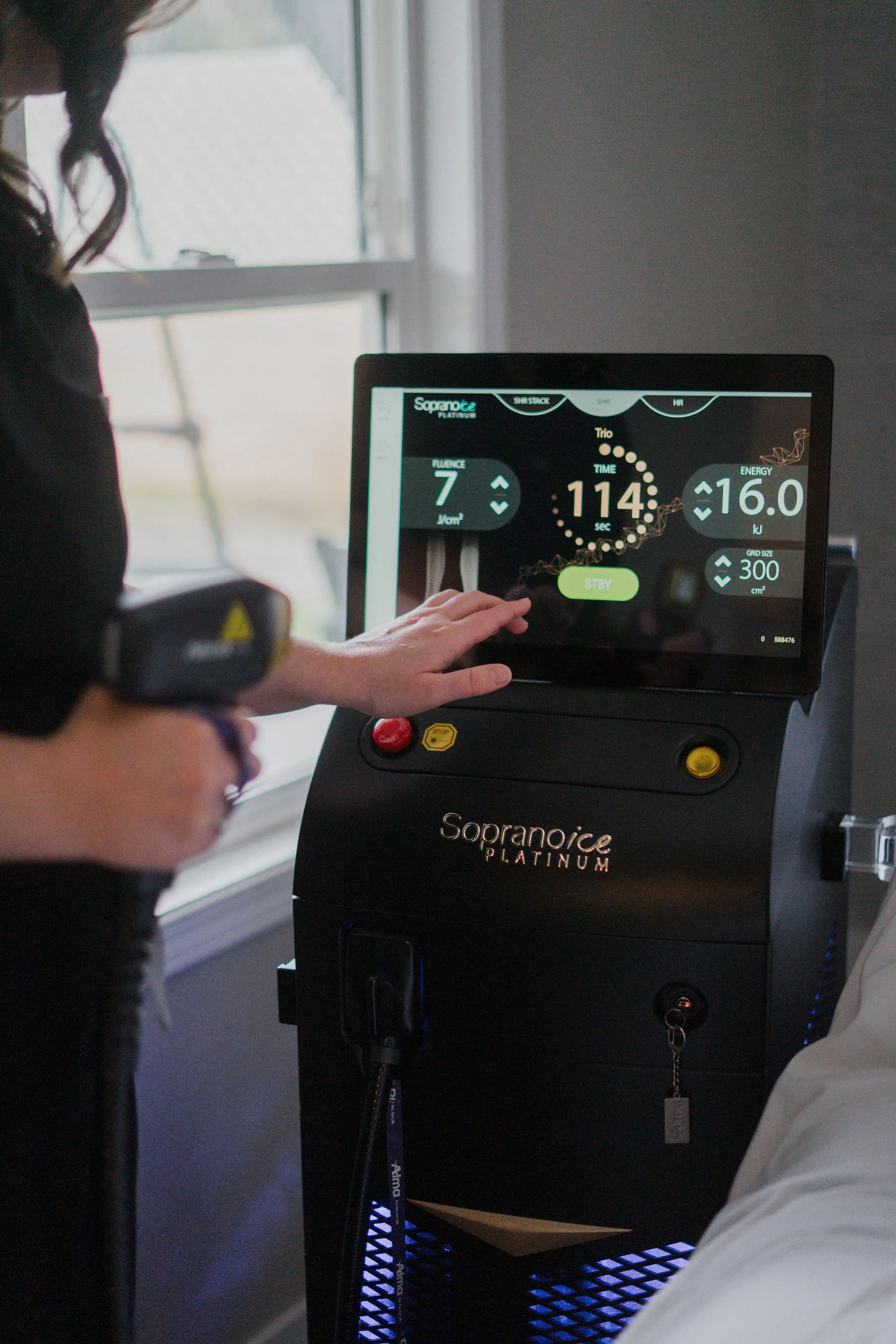 A person operating a Soprano Ice Platinum laser device, used for hair removal, with a large display screen showing settings such as time, energy level, and categories like SHR, HR, and SHR STACK. The device is black with branding on the front.