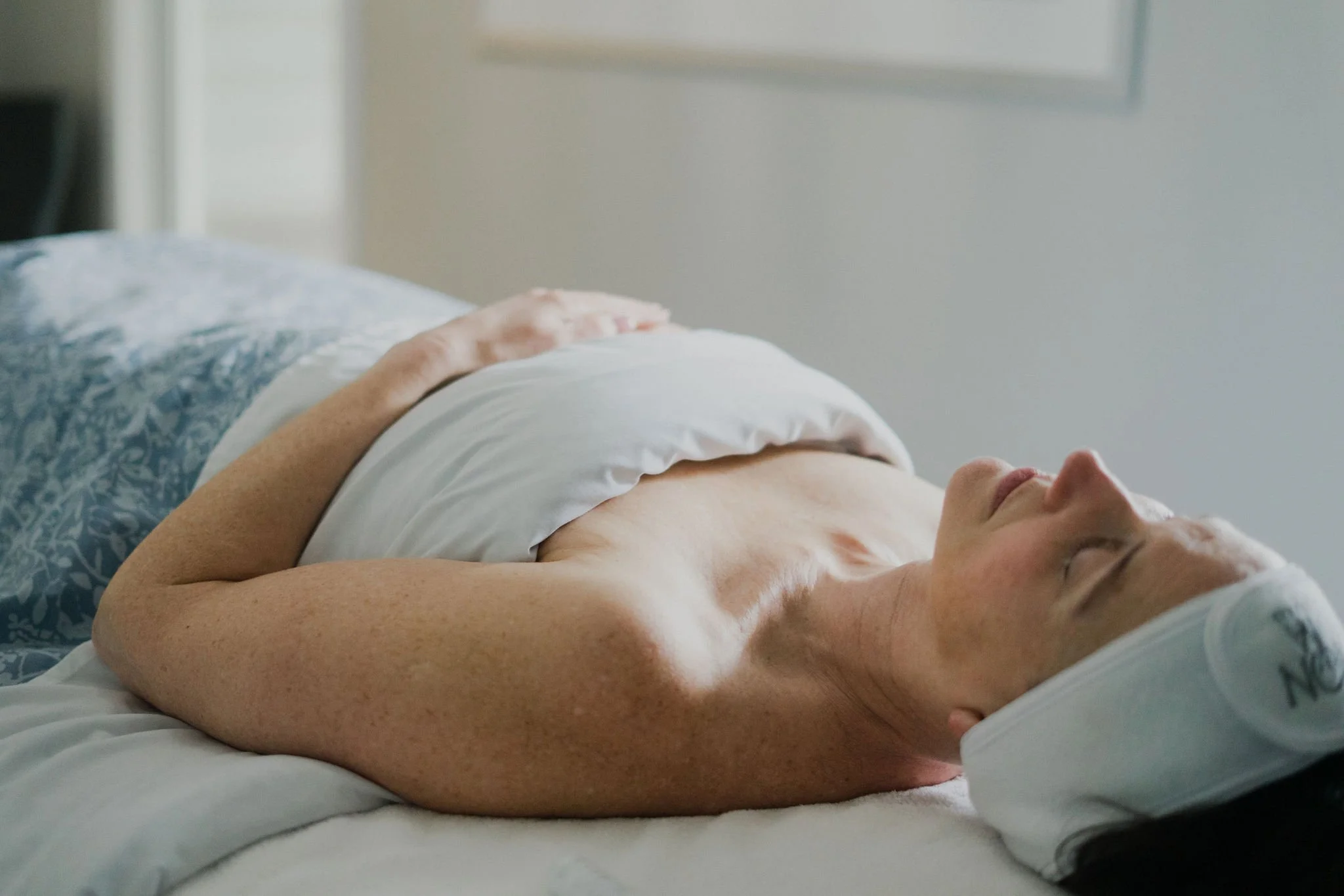 A person lying down on a bed with their eyes closed, resting with one hand on their abdomen, in a bedroom with a light-colored wall.