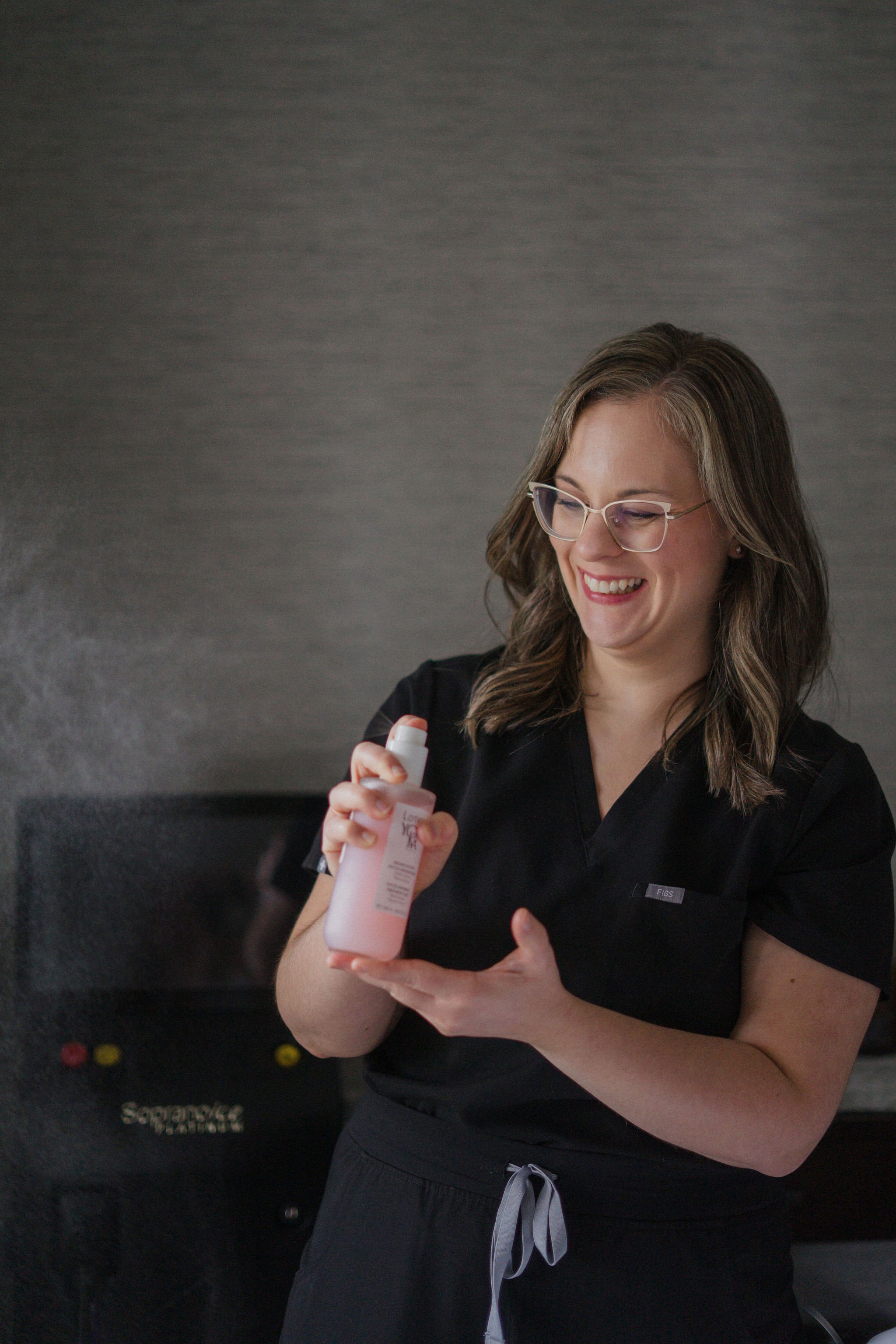 A woman with glasses and brown hair is smiling and holding a pink bottle of moisturizer or lotion, wearing a black top, standing in front of a gray wall.