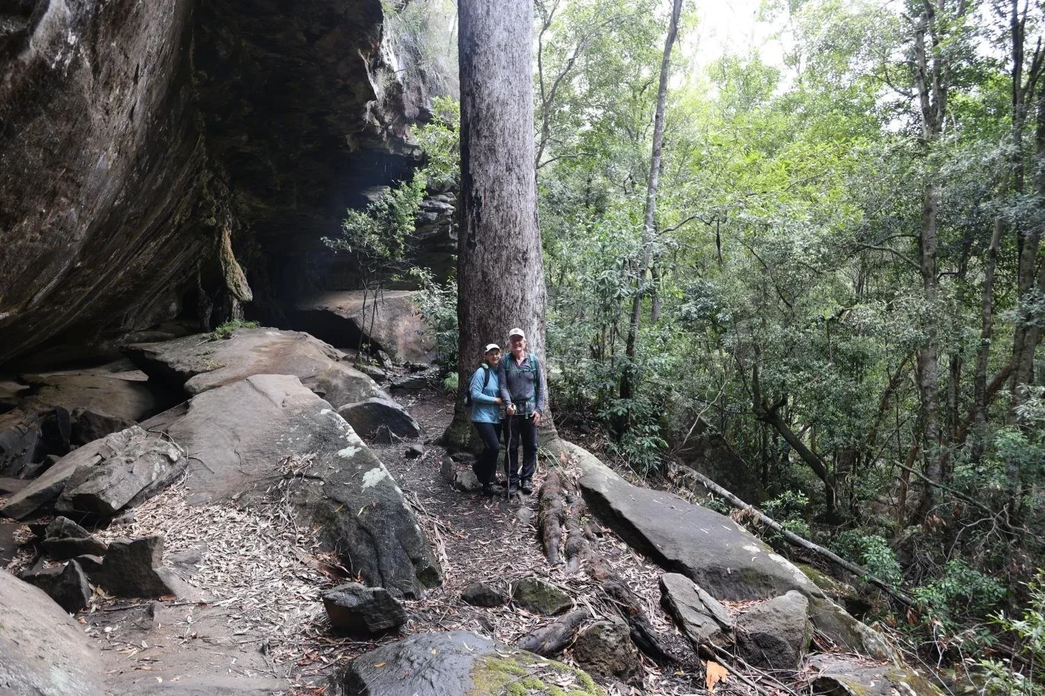 Two hikers standing beside a large sandstone rock overhang at Palona Cave in the Royal National Park, surrounded by eucalypt forest