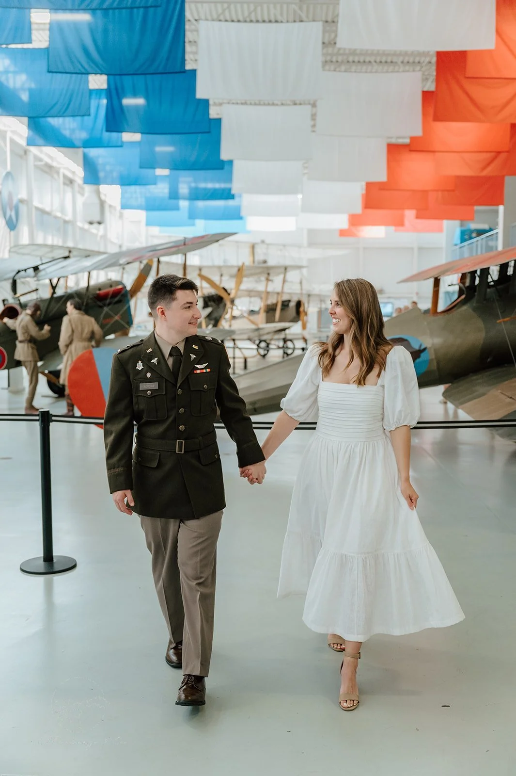 A couple holding hands and walking inside an aviation museum, with vintage airplanes displayed in the background and colorful fabric flags hanging from the ceiling.