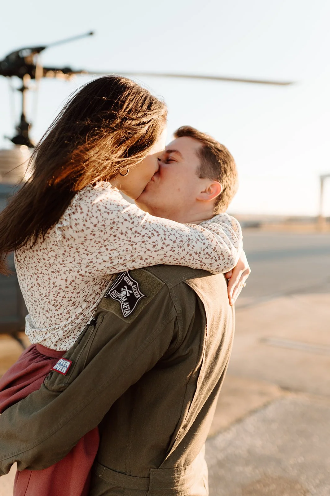 A woman and a man are sharing a kiss outdoors, with the woman being carried by the man in a joyful embrace. The scene is set during sunset with a helicopter visible in the background.