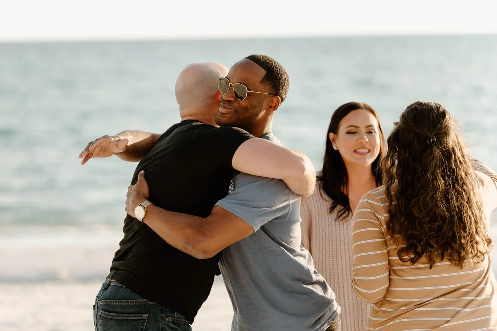  Two men hug in congratulations after one proposed to his fiancee on a beach 