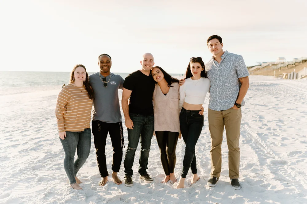  A happy newly engaged couple stand on a beach with family and friends smiling at sunset 