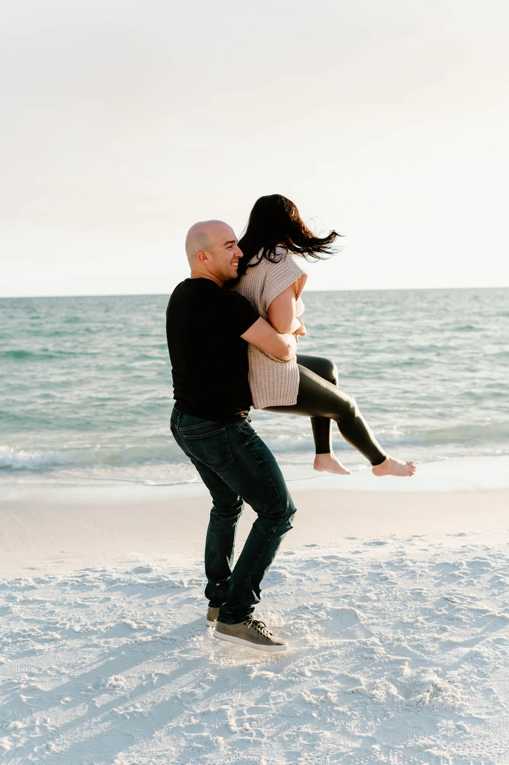  A man lifts and spins his fiancee on a beach in jeans and a black shirt 