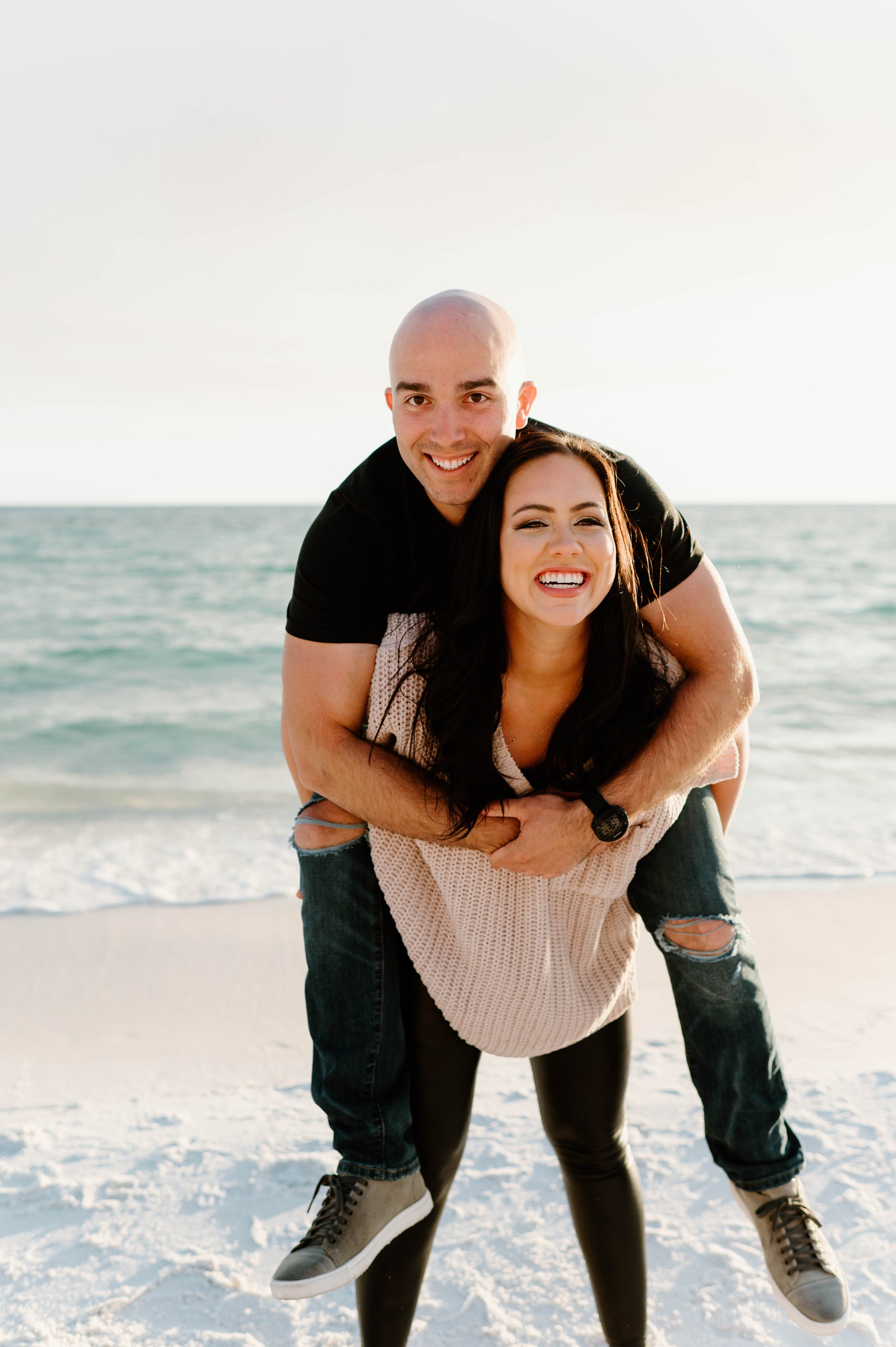  A man climbs on his fiancee's back while they walk on a beach at sunset 