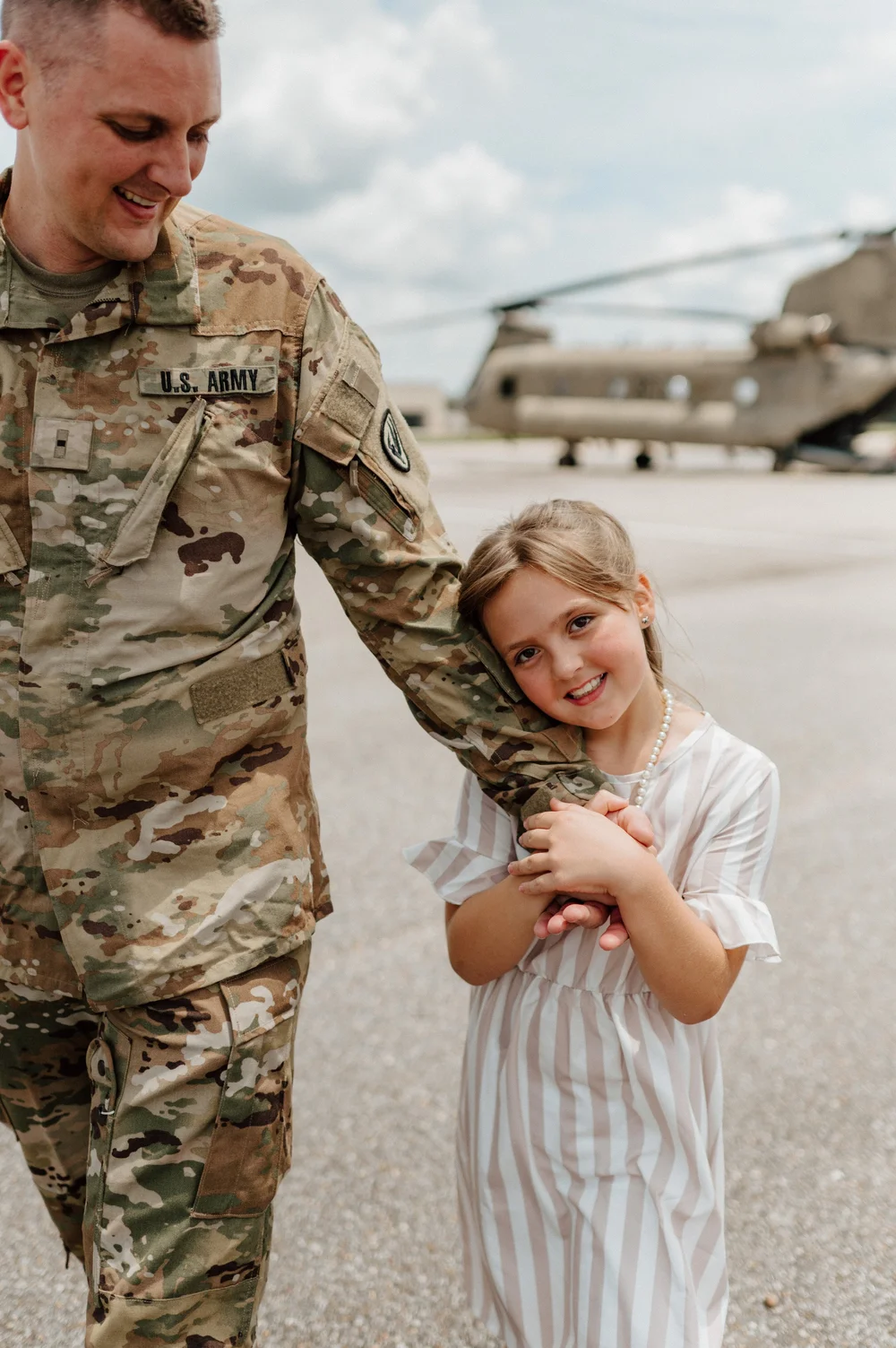  A toddler girl in a stripe dress hangs on the hand on her dad after his Military Graduation 