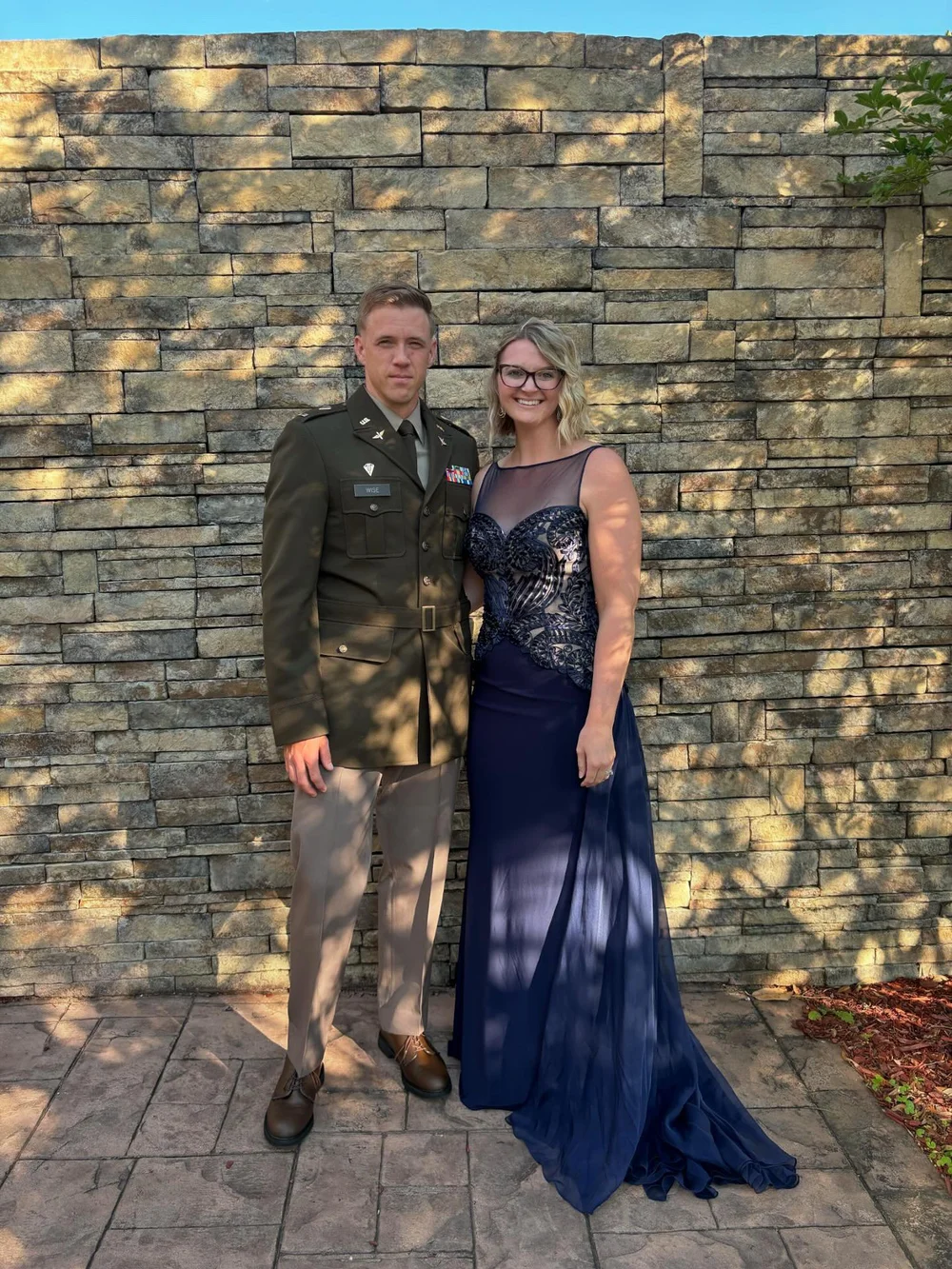  A happy couple stands against a stone wall smiling and holding hands 