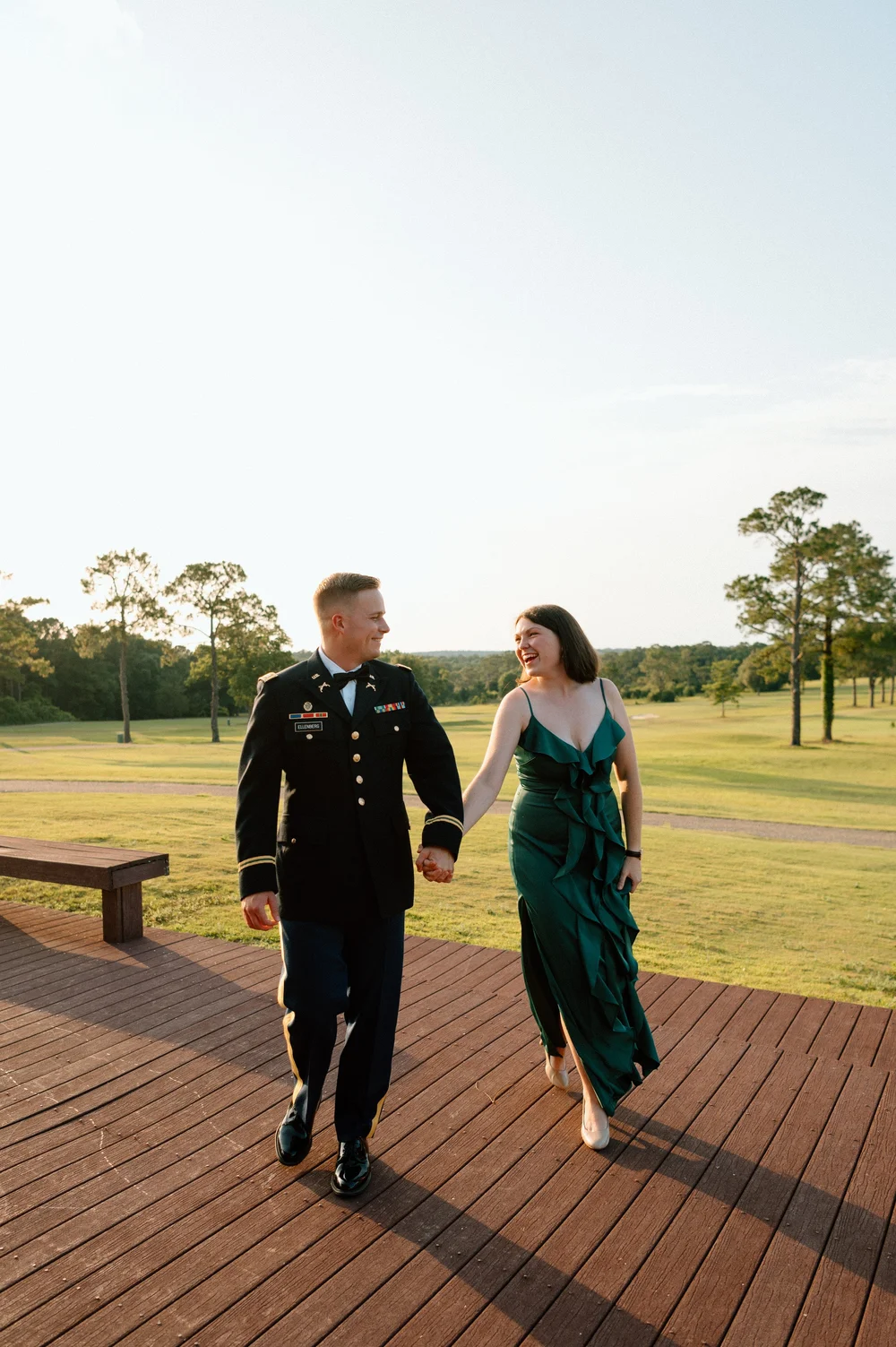  A happy couple walks hand in hand on a deck at sunset while smiling at each other 