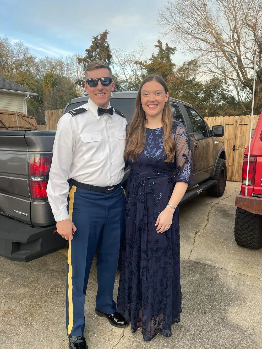  A man in sunglasses stands with his partner in front of a truck while smiling big 
