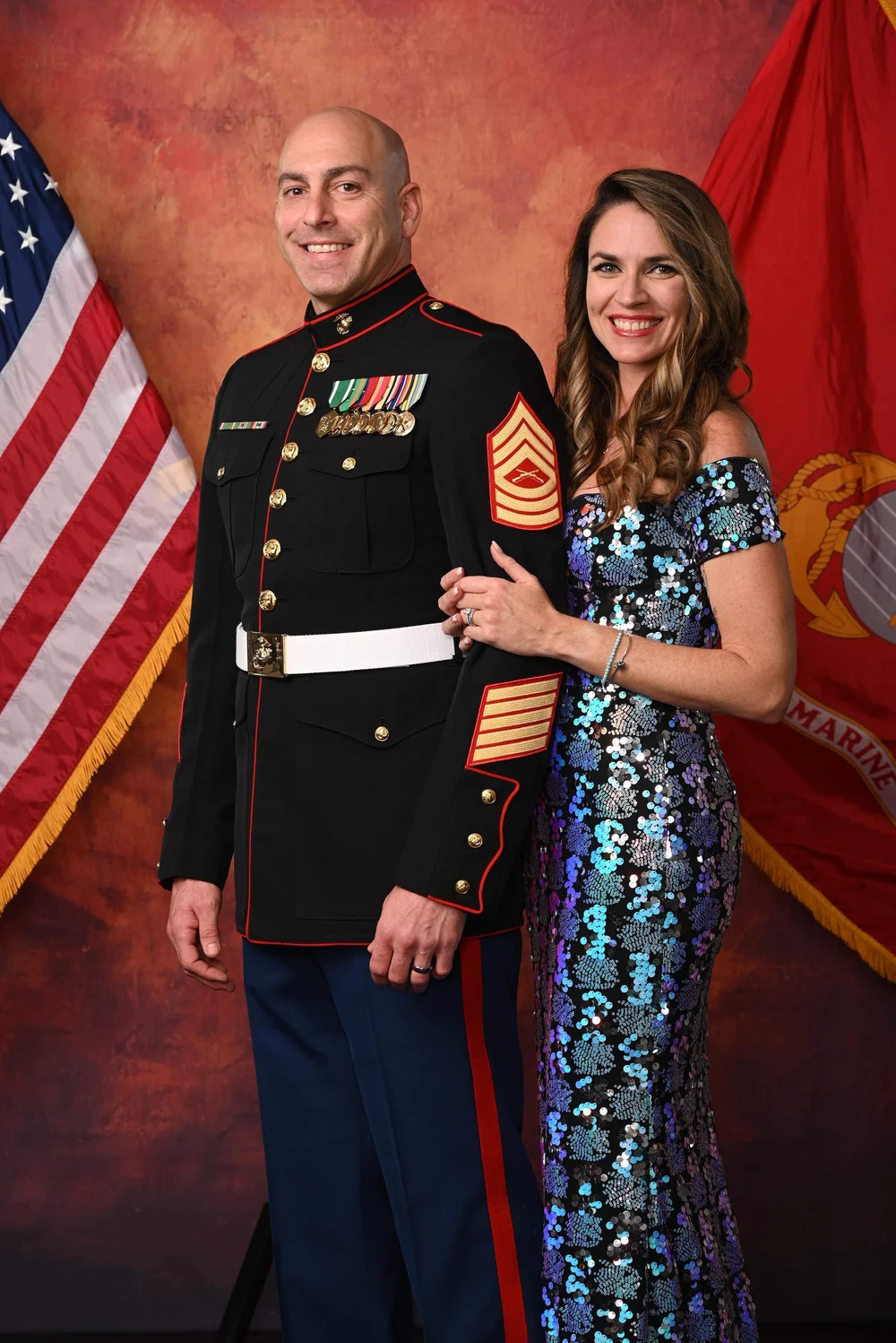  A decorated officer stands by a flag while his wife hangs on his arm in a blue dress to celebrate his Military Graduation 