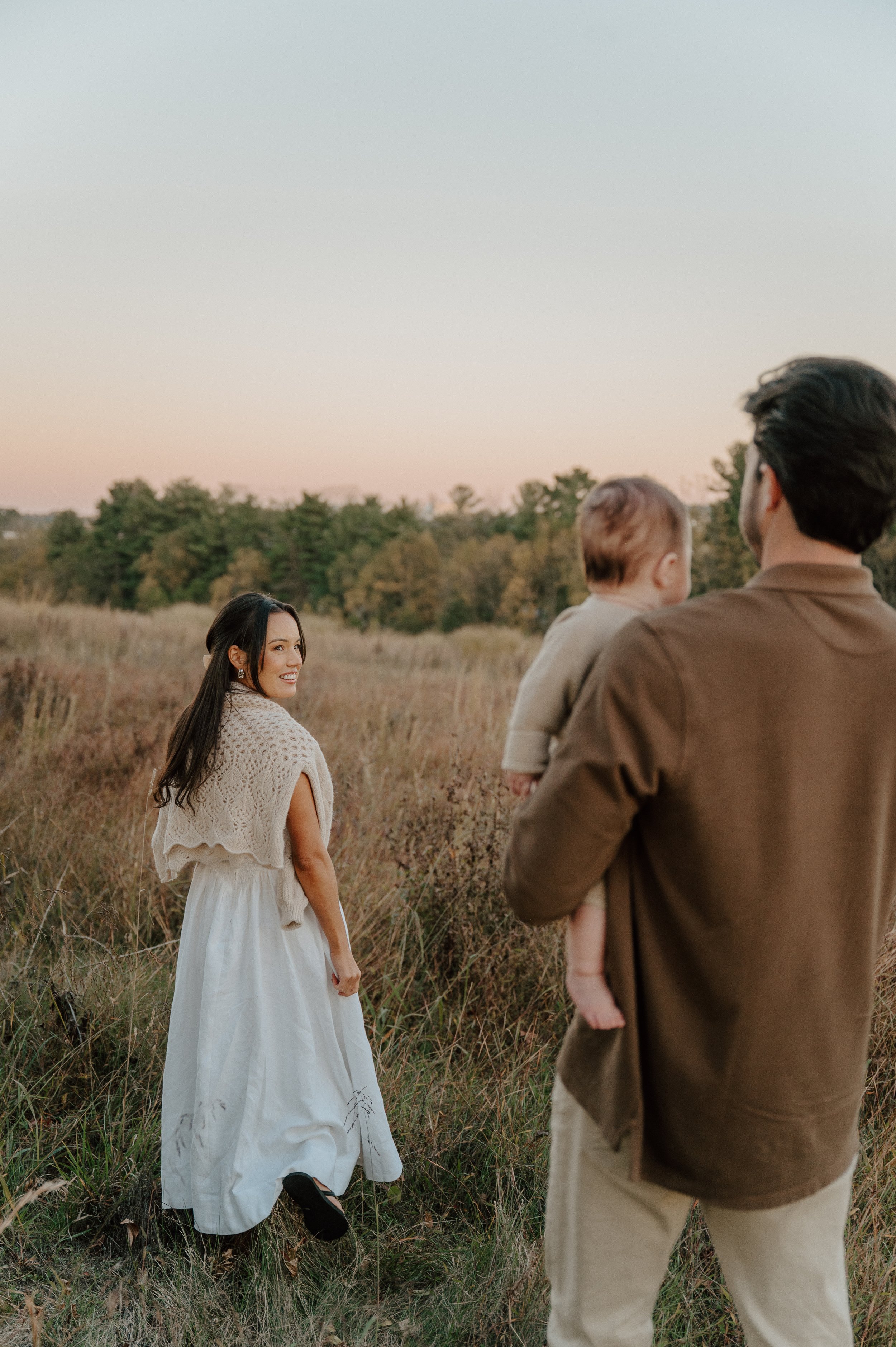 A woman in a white dress and beige shawl stands in a field during sunset, facing a man holding a child, with trees in the background.