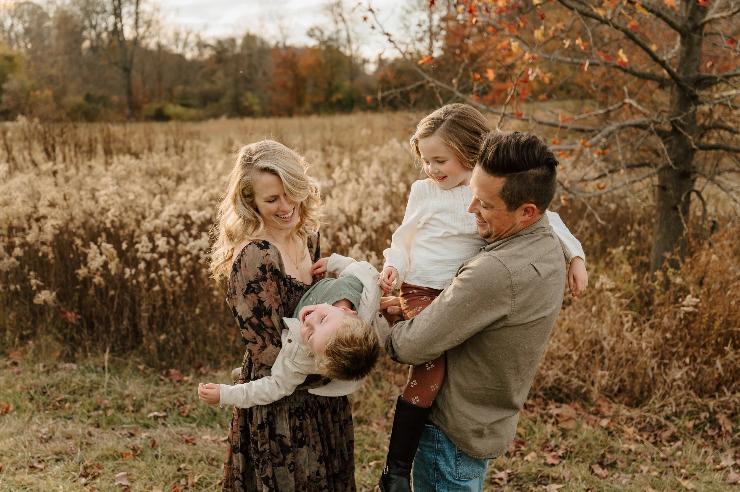A family of four playing outdoors in an autumn setting with trees and grasses around, the mother and father holding and laughing with two children.