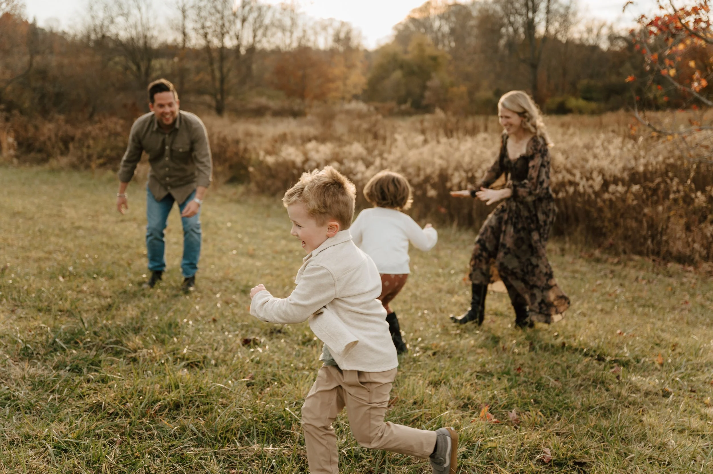 A family of four, including two young children, playing and running in a grassy field during autumn with trees and brush in the background.