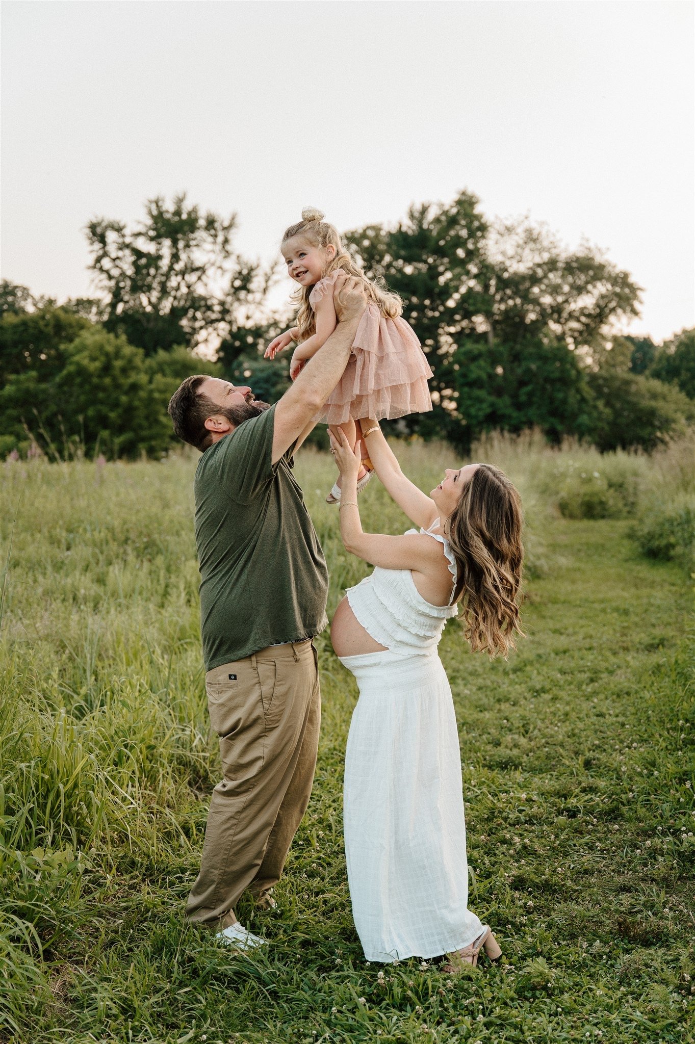 A family of three outdoors, with a father lifting a young girl in a pink dress, and a mother supporting her, standing on grass with trees in the background.