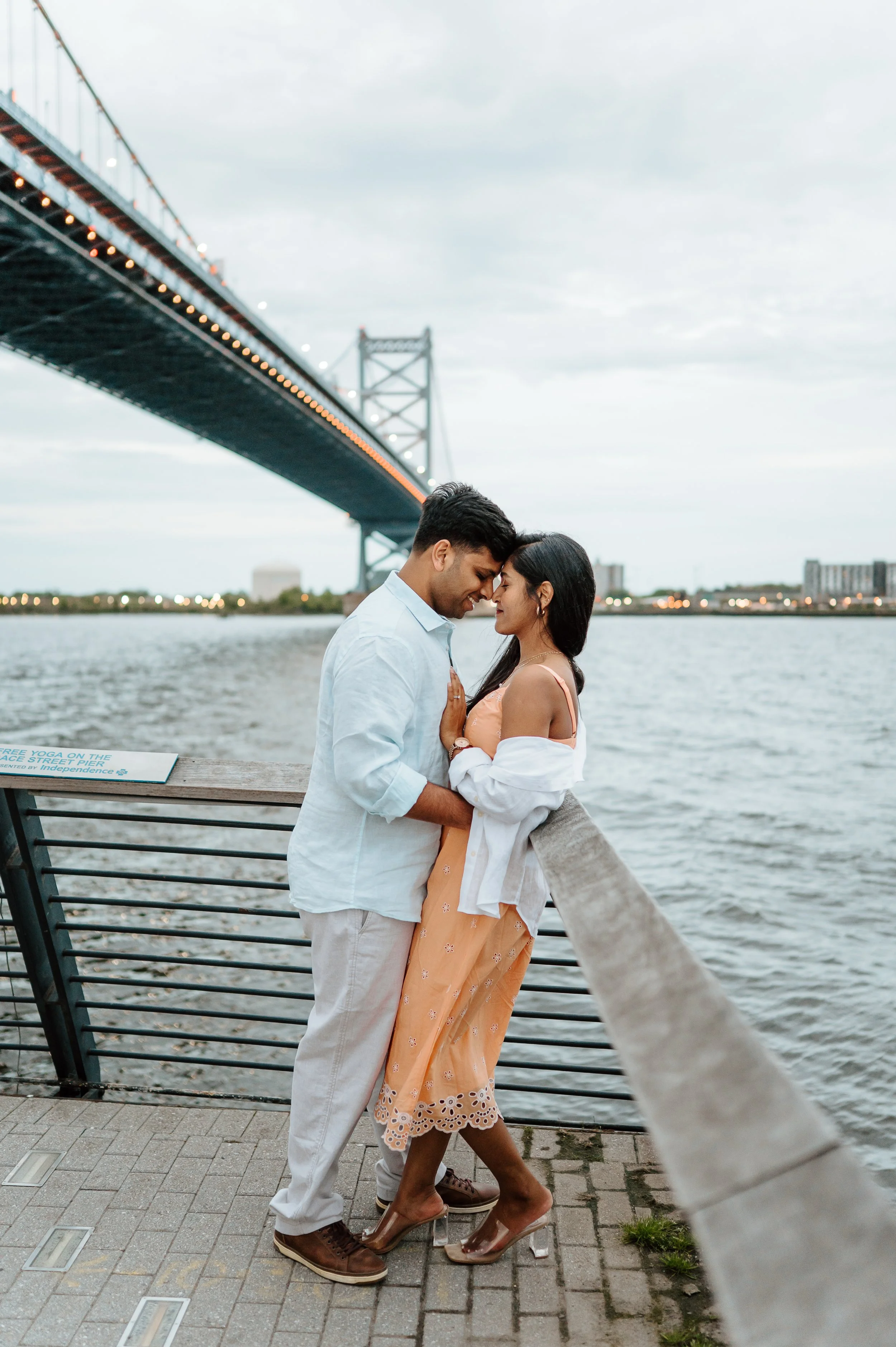 A tender moment shared by a man and woman on a waterfront, with a bridge in the background under an overcast sky.