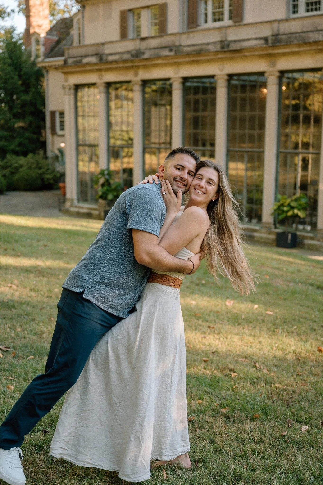 A couple happily embraces outdoors in front of a large glass house with potted plants, during daylight.