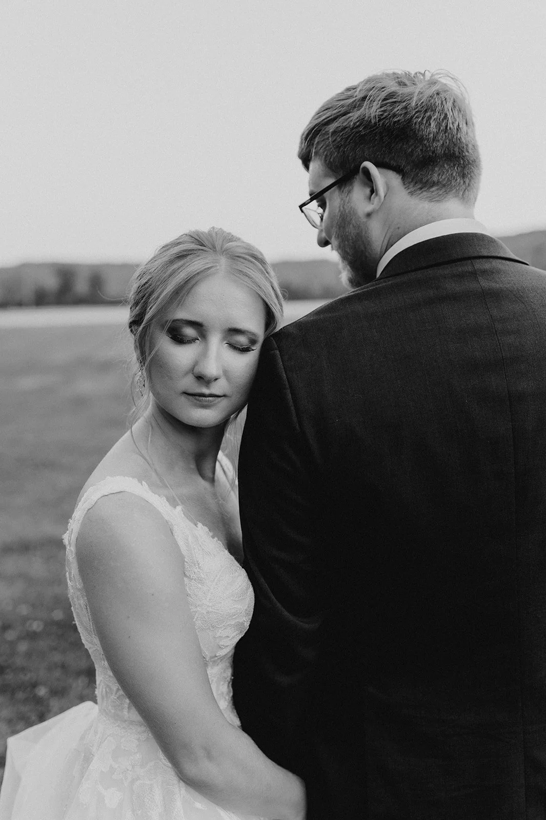 Black and white photo of a bride and groom on their wedding day, embracing outdoors. The bride has her eyes closed and is leaning her head on the groom's chest; the groom has glasses and is looking down at her. The background shows a field with distant hills.