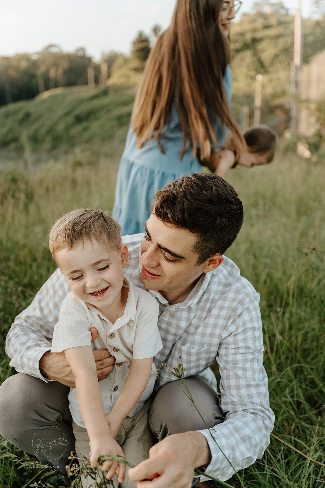 A man and young boy sitting on the grass, smiling and playing with a plant, outdoors in a grassy field. A woman and child are visible in the background.
