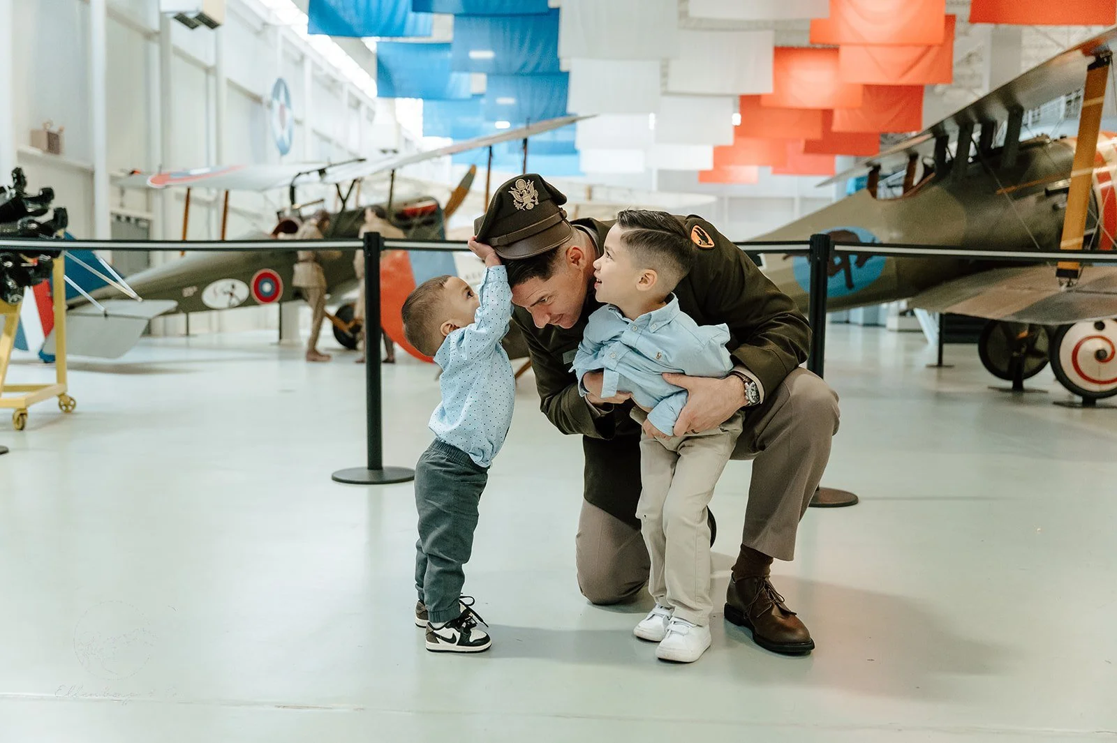 A navy officer kneeling and playing with two young boys in an aviation museum, with vintage airplanes in the background.