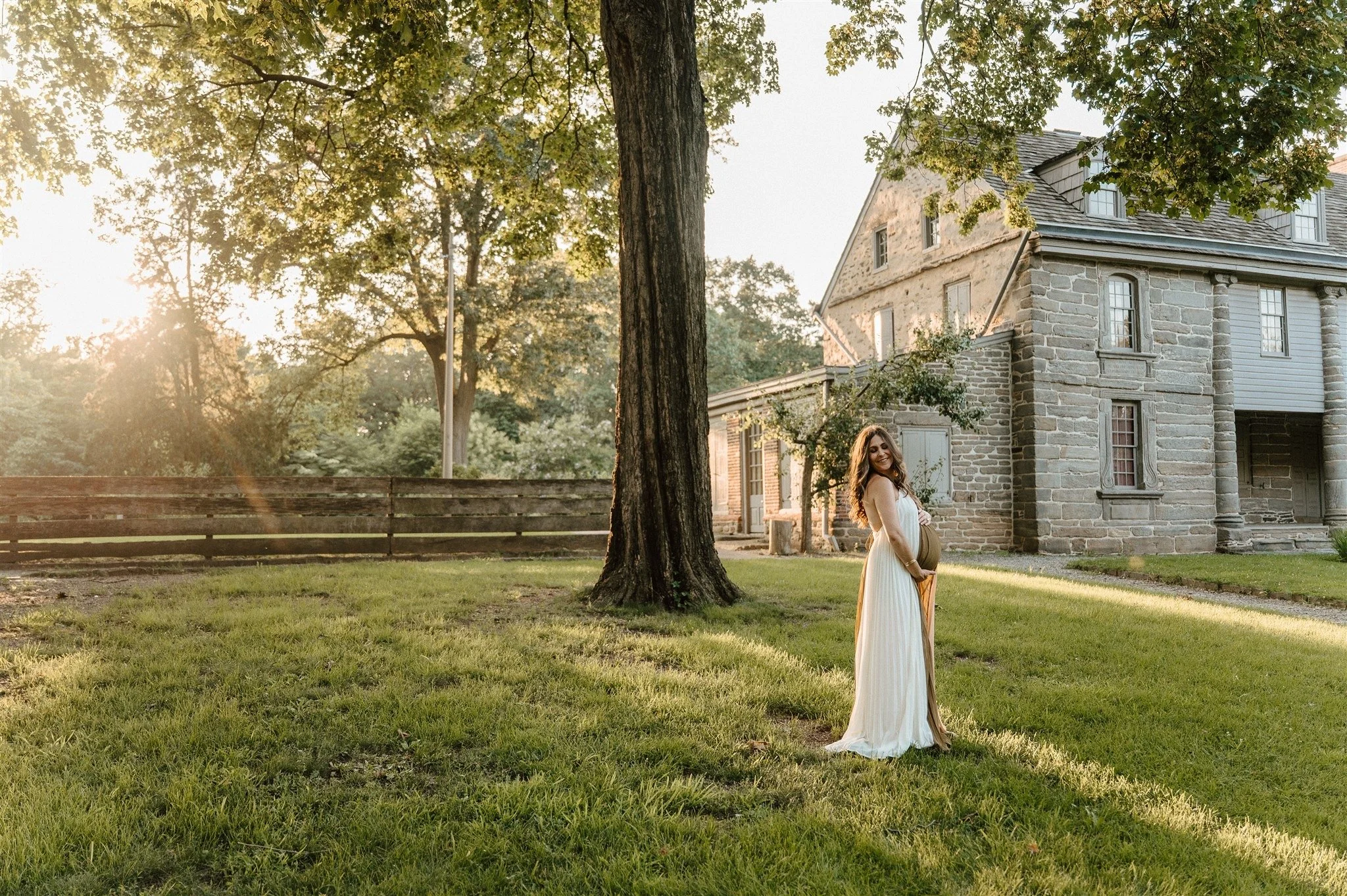 A pregnant woman in a long white dress standing outdoors on a grassy lawn near a large tree, with a stone building in the background, illuminated by warm sunlight at sunset.