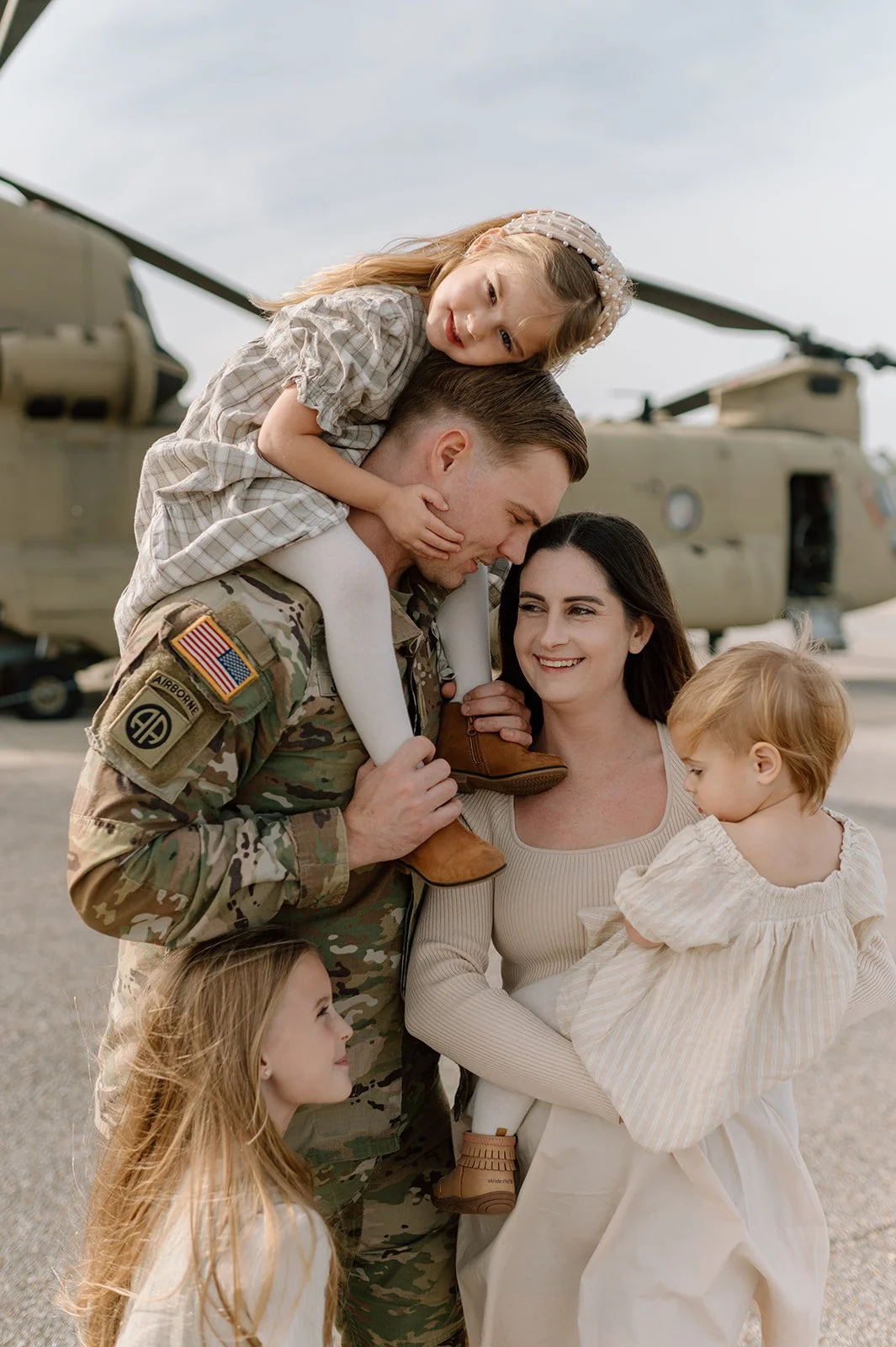 A soldier in camouflage uniform carrying a young girl on his shoulders, smiling at her. A woman smiling at the soldier holds a young girl in her arms. A helicopter is in the background on a tarmac.