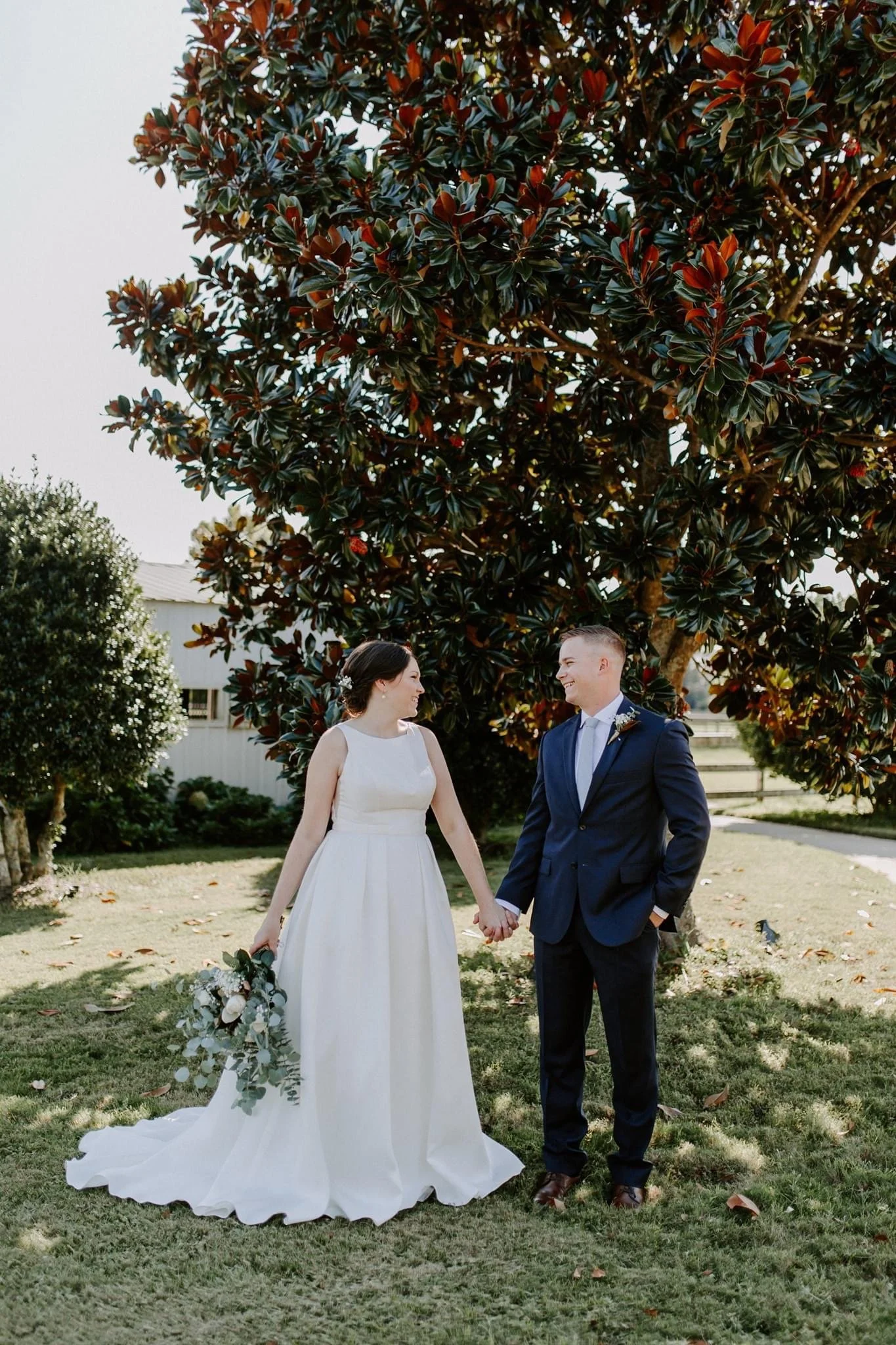 A bride and groom holding hands outdoors under a large tree, smiling at each other. The bride wears a white wedding dress and holds a bouquet; the groom is dressed in a navy suit with a tie. The scene is sunny, with green grass and some fallen leaves.