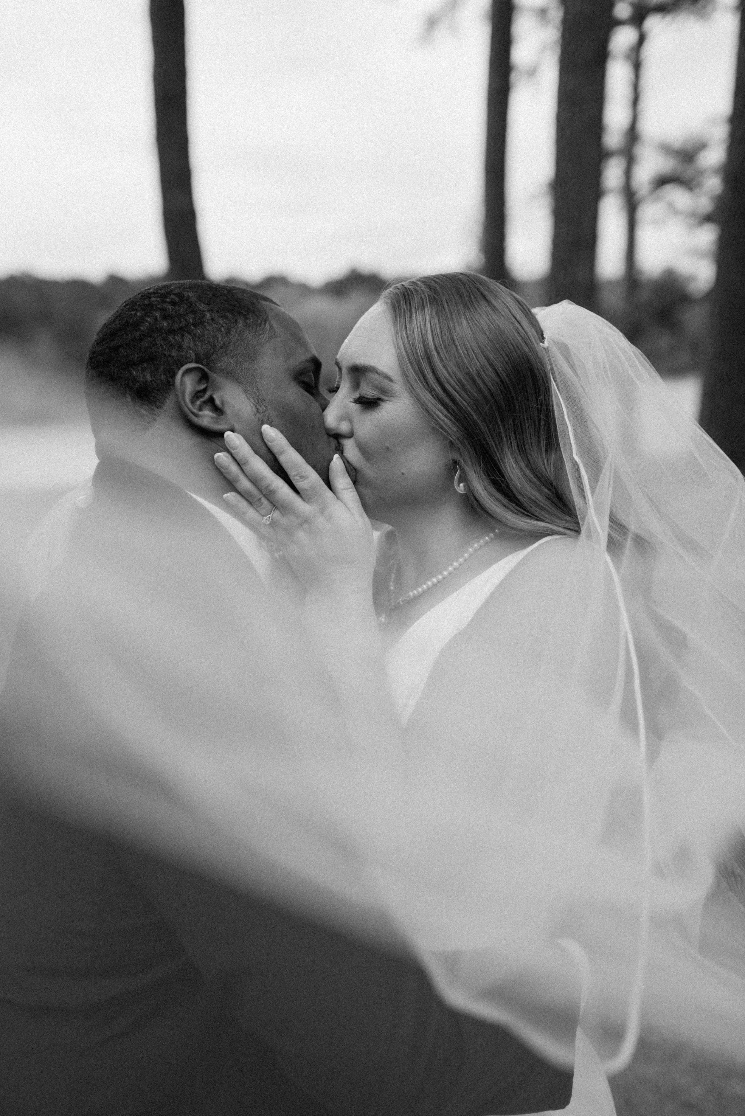 A black-and-white photo of a bride and groom sharing a kiss outdoors, with trees in the background.