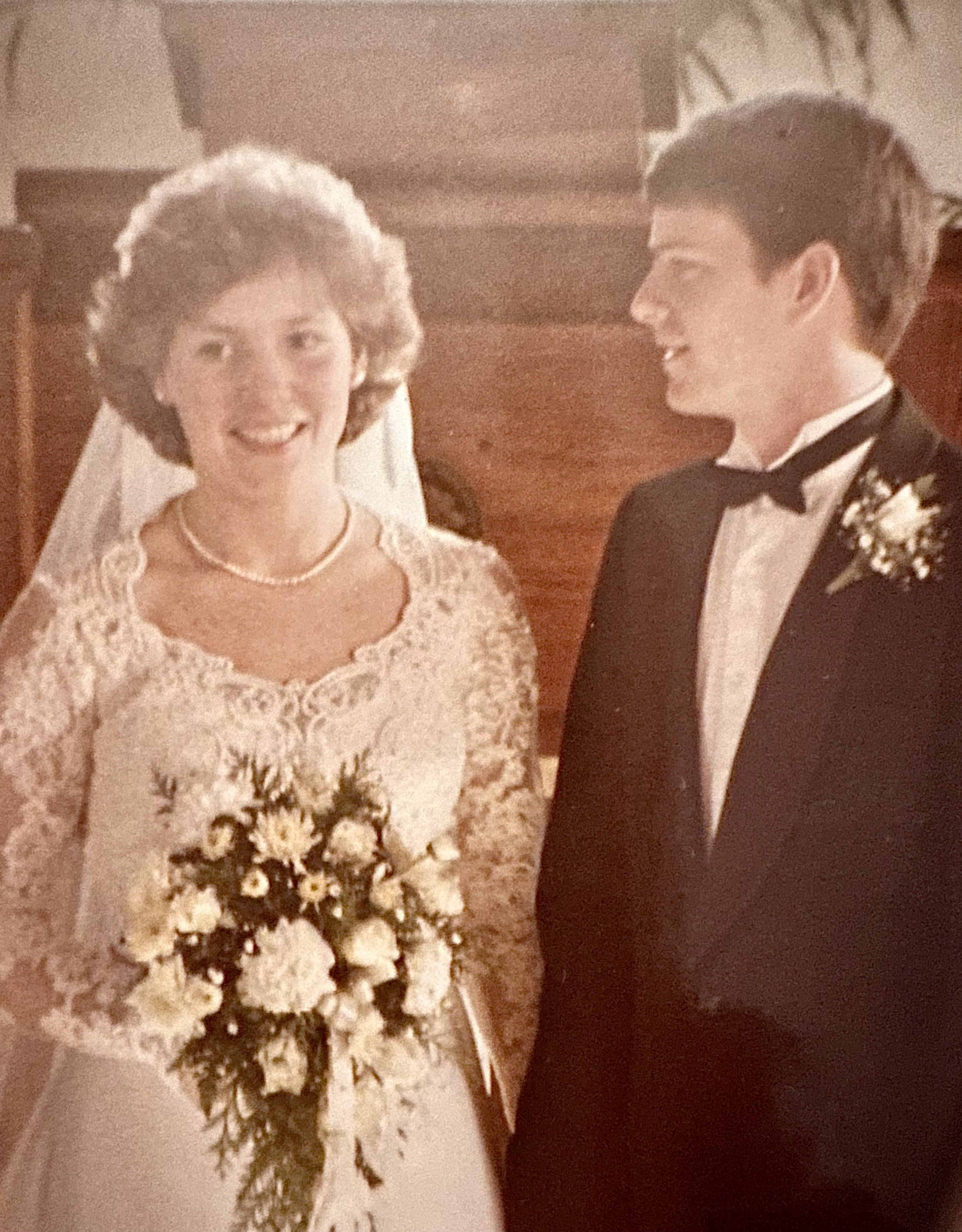 A bride in a lace wedding dress holding a bouquet of white flowers, smiling, standing next to a groom in a tuxedo with a boutonnière, both inside a church.