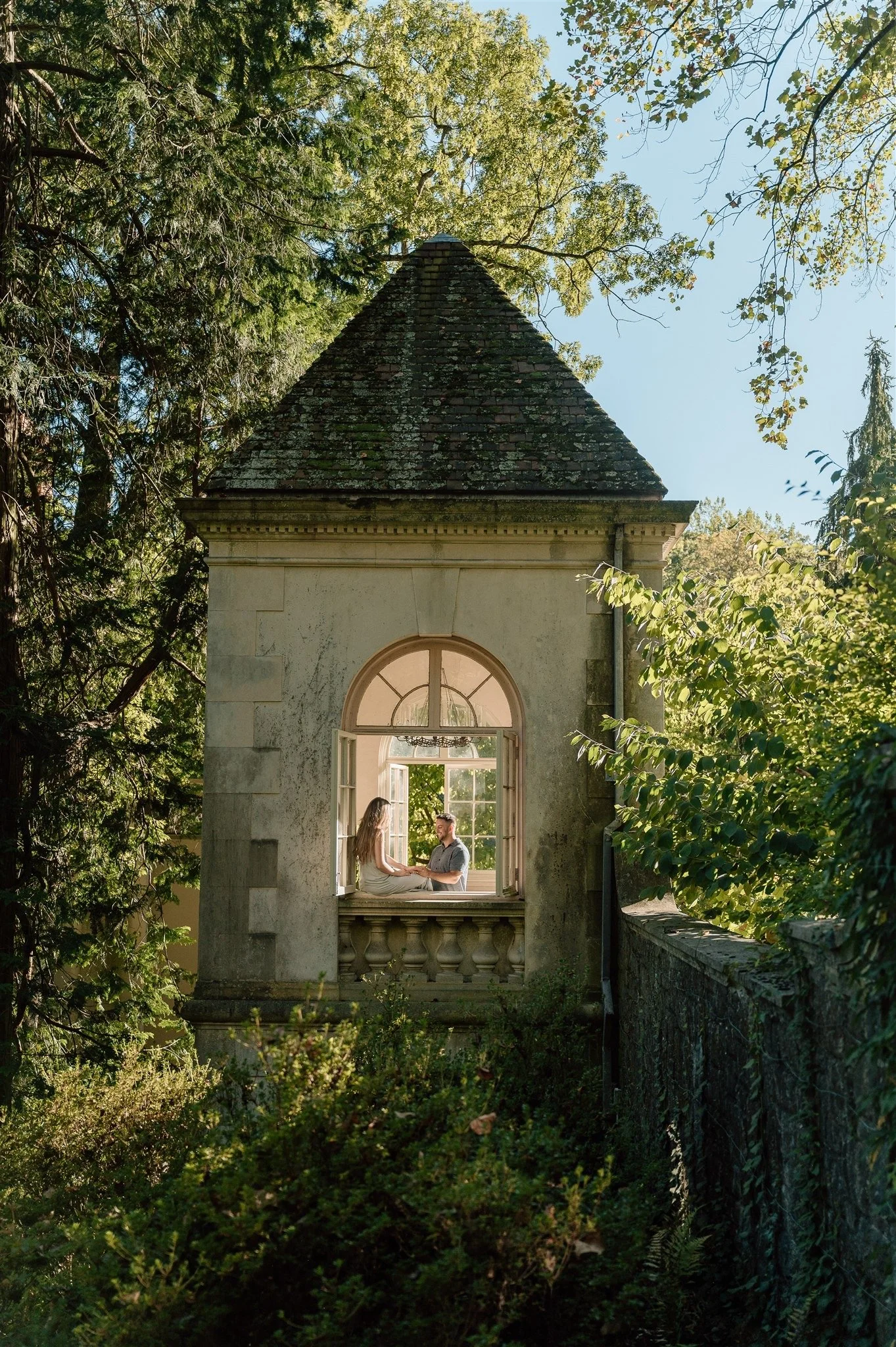 A couple sitting on a windowsill inside a small, historic building surrounded by trees and greenery.