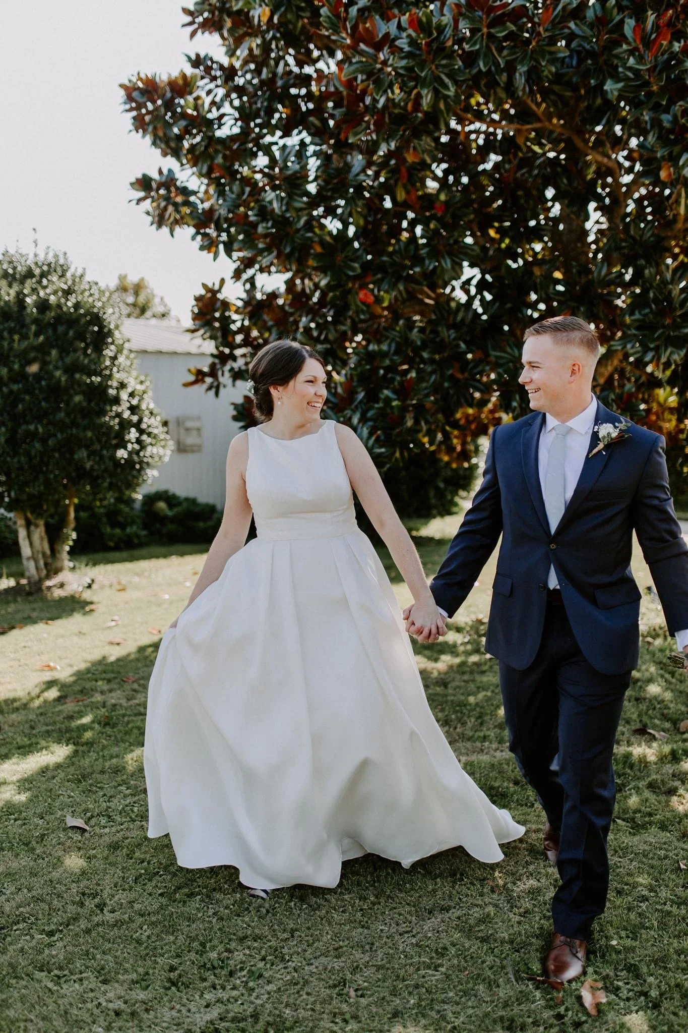 A bride and groom holding hands and walking outdoors on a sunny day, smiling at each other, with trees and a white building in the background.