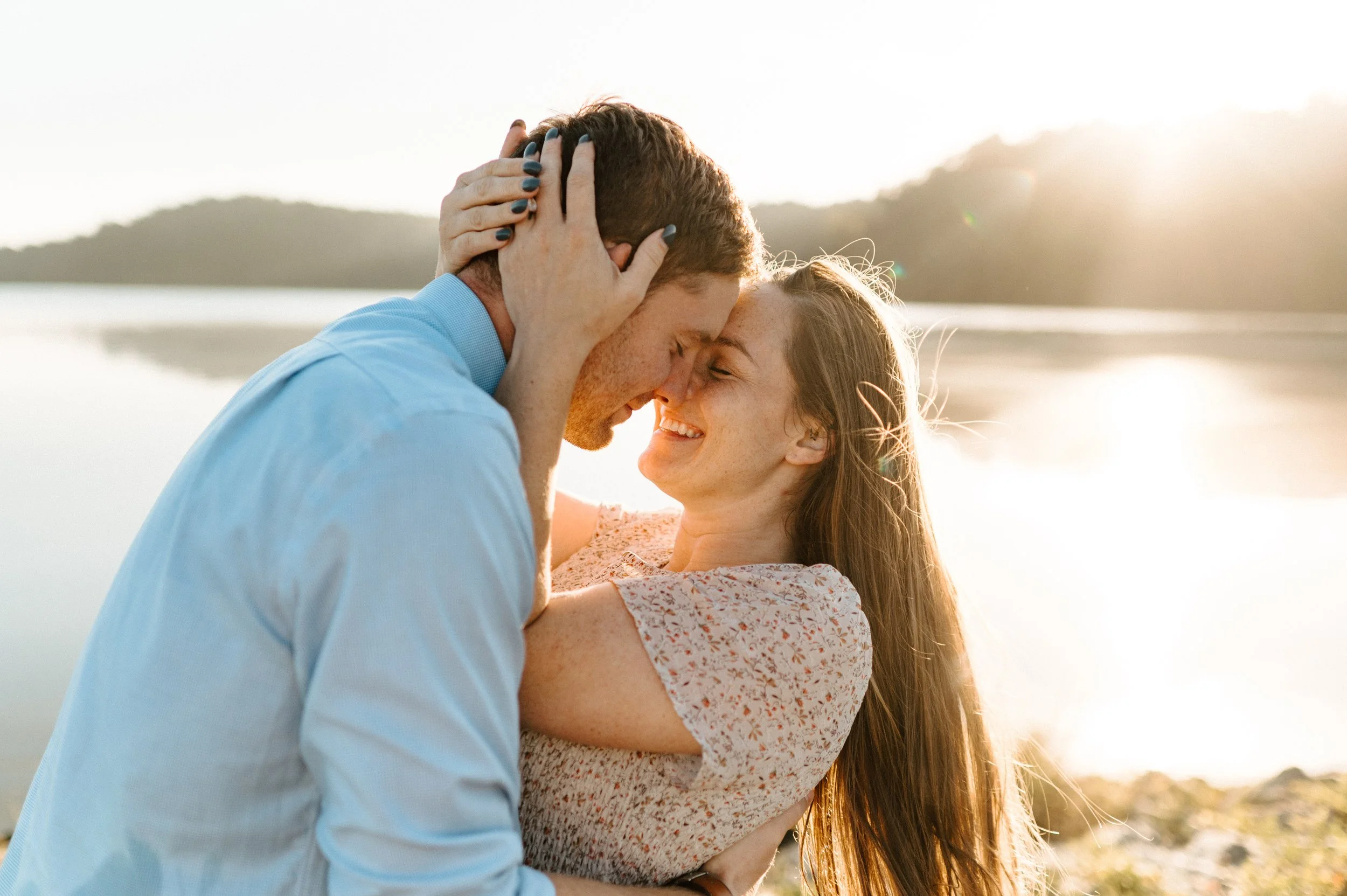 A couple embracing and smiling near a body of water during sunset.