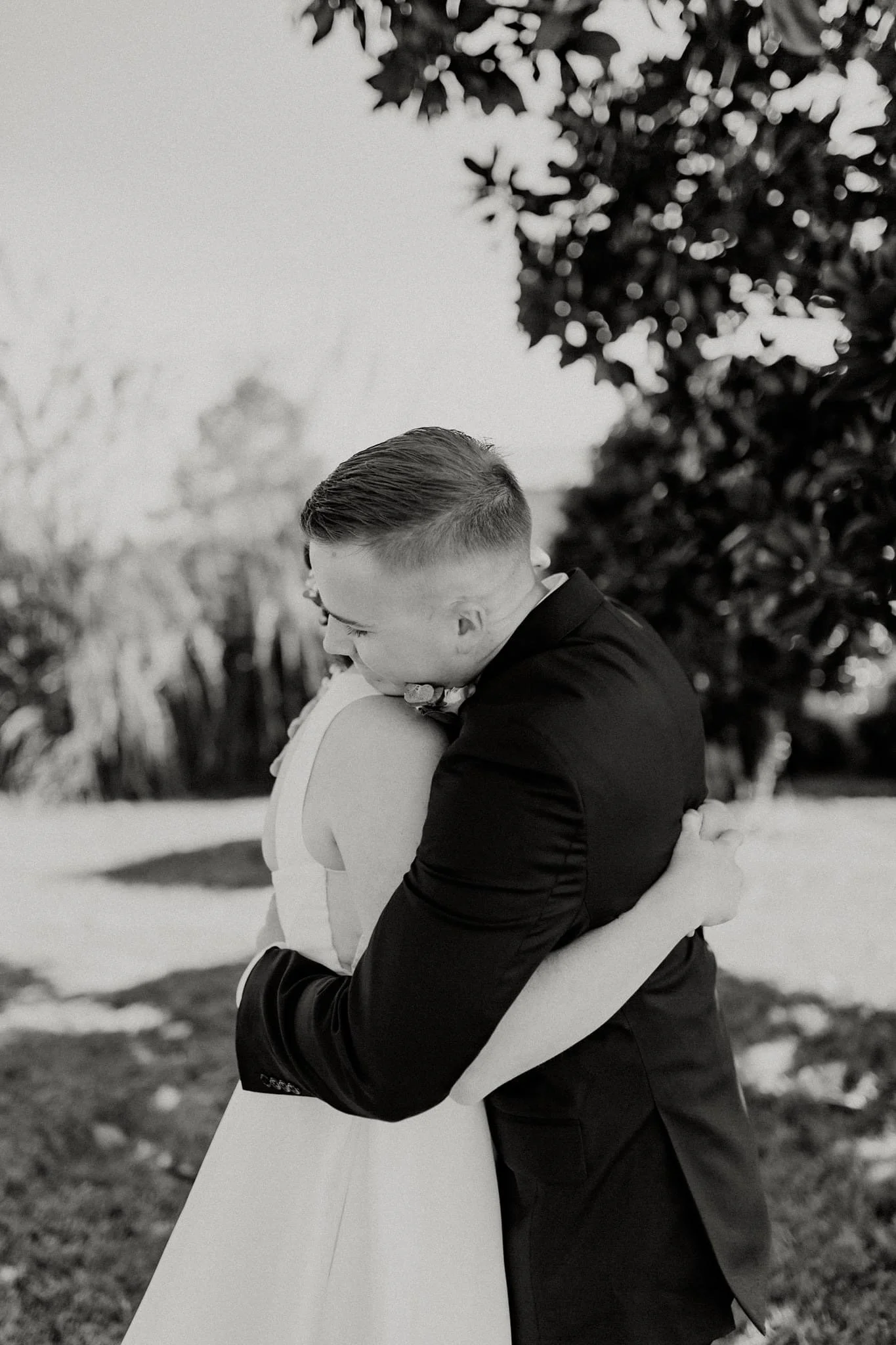 A black and white photo of a man and woman embracing outdoors, likely at a wedding or special event, with trees in the background.