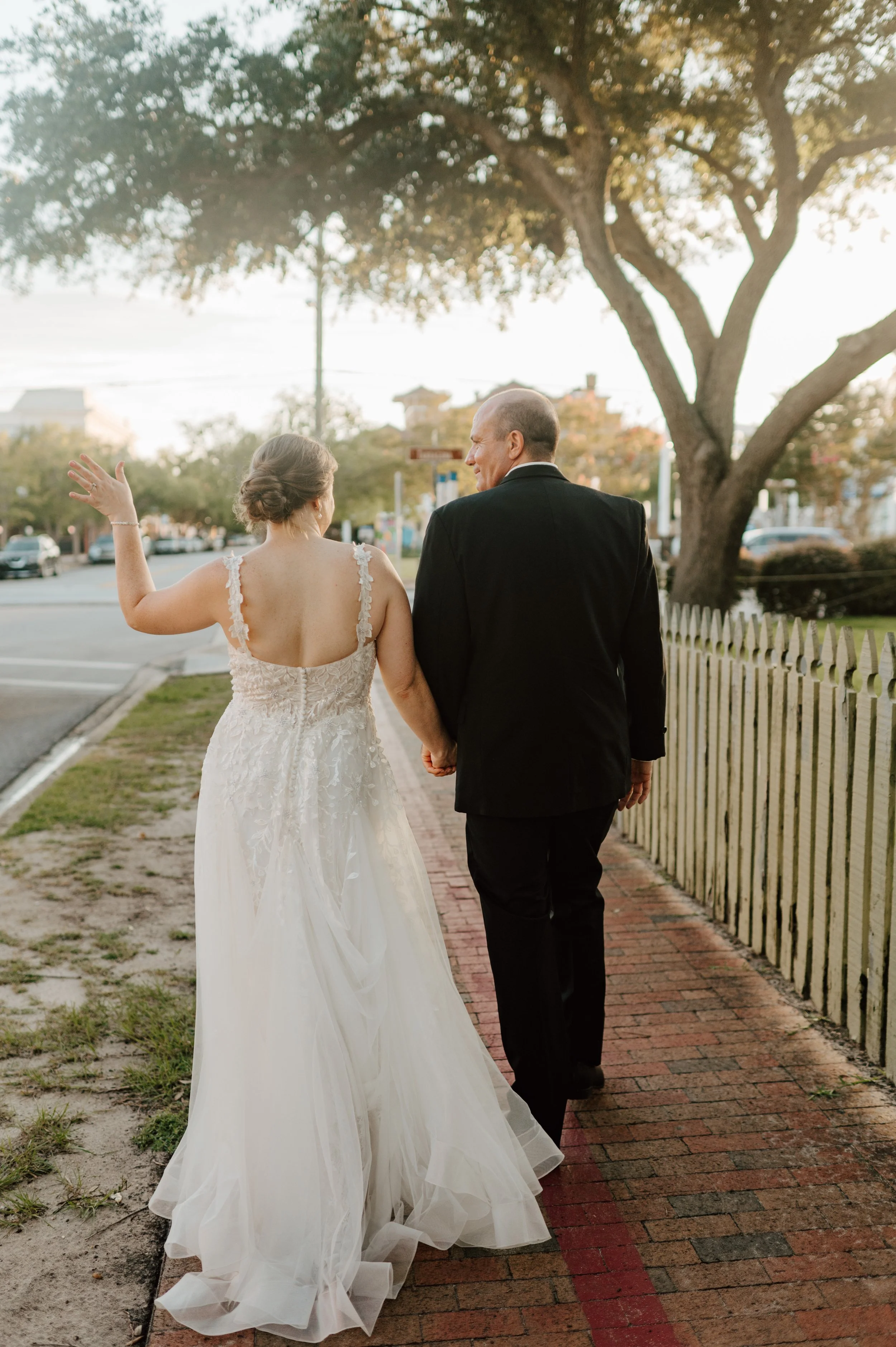 A bride and groom walking hand in hand down a brick sidewalk, holding hands, with a white picket fence on the side and trees in the background, during sunset.