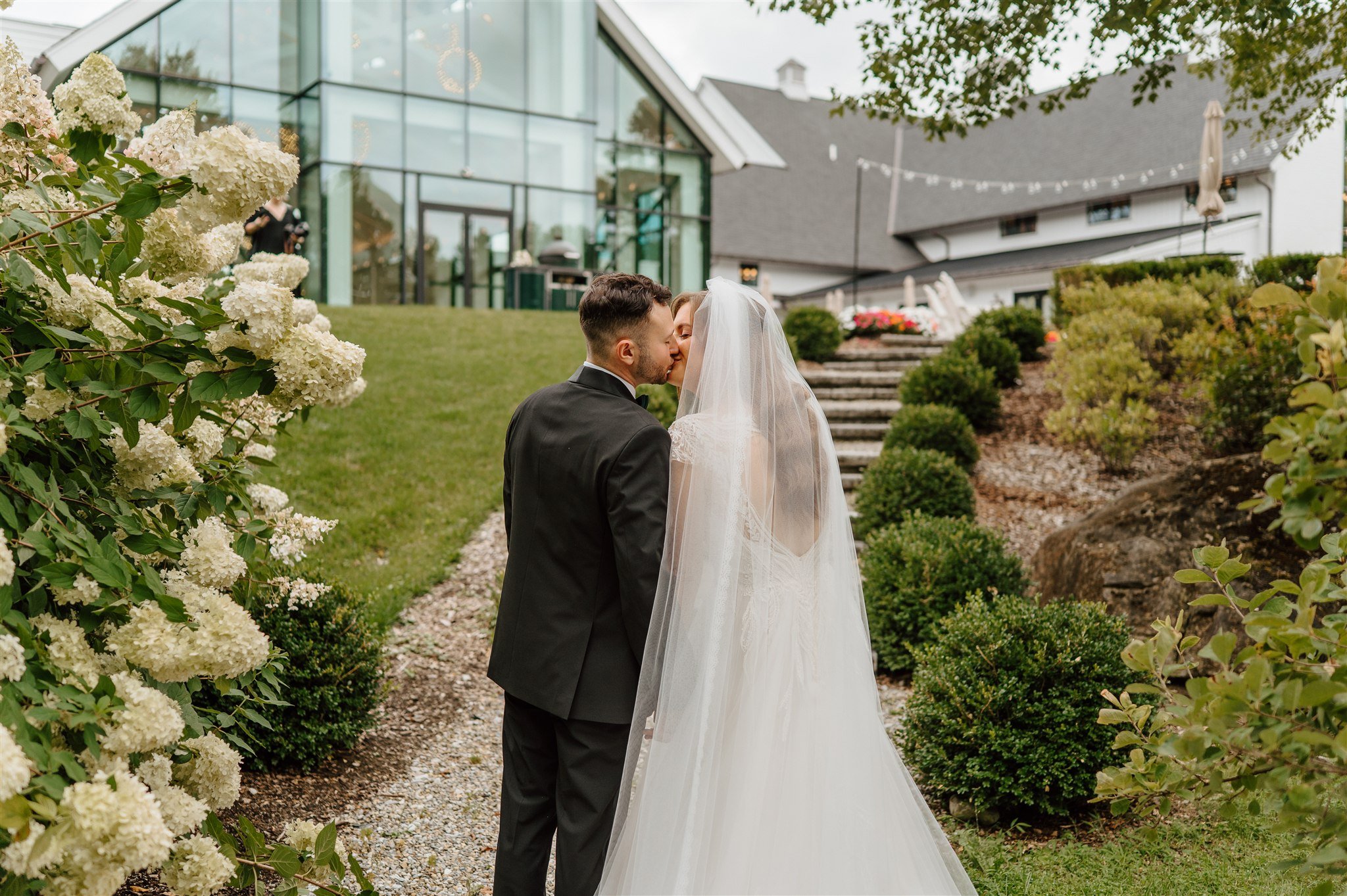 A newly married couple sharing a kiss outdoors, with the bride in a white wedding gown and veil, and the groom in a black suit, surrounded by green bushes and blooming hydrangeas, in front of a modern glass building and a house with string lights.