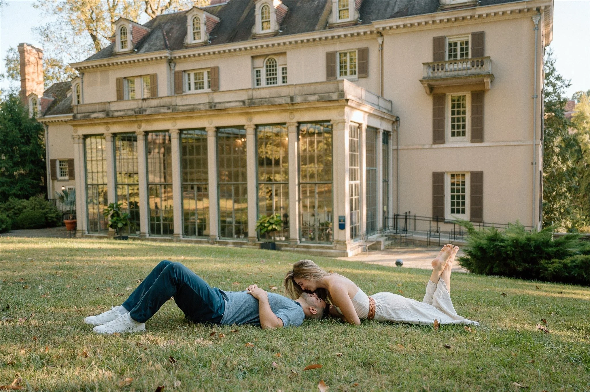 A couple lying on the grass in front of a large, historic house. The woman is leaning over the man, and they are smiling at each other, enjoying a tender moment outdoors.