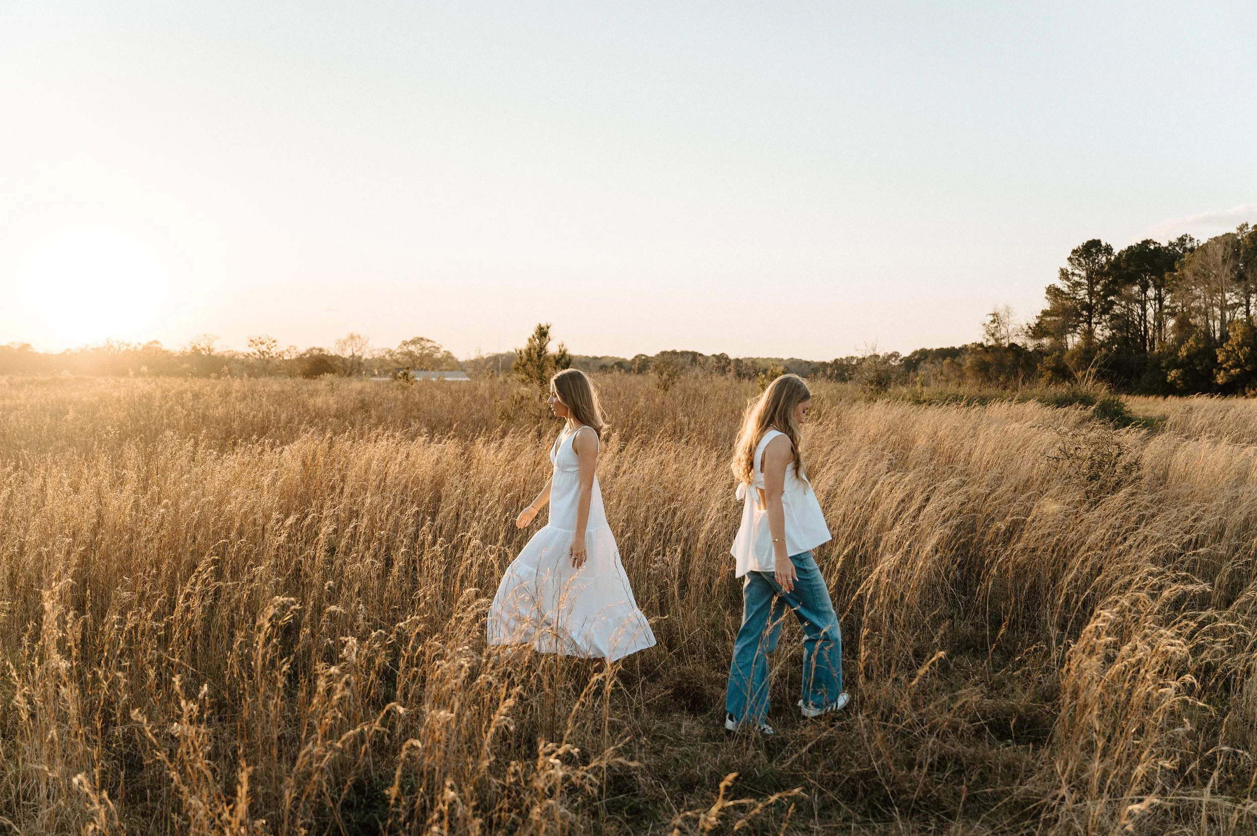 Two women walk through a golden field of tall grass at sunset, one in a white dress and the other in a white top and jeans.