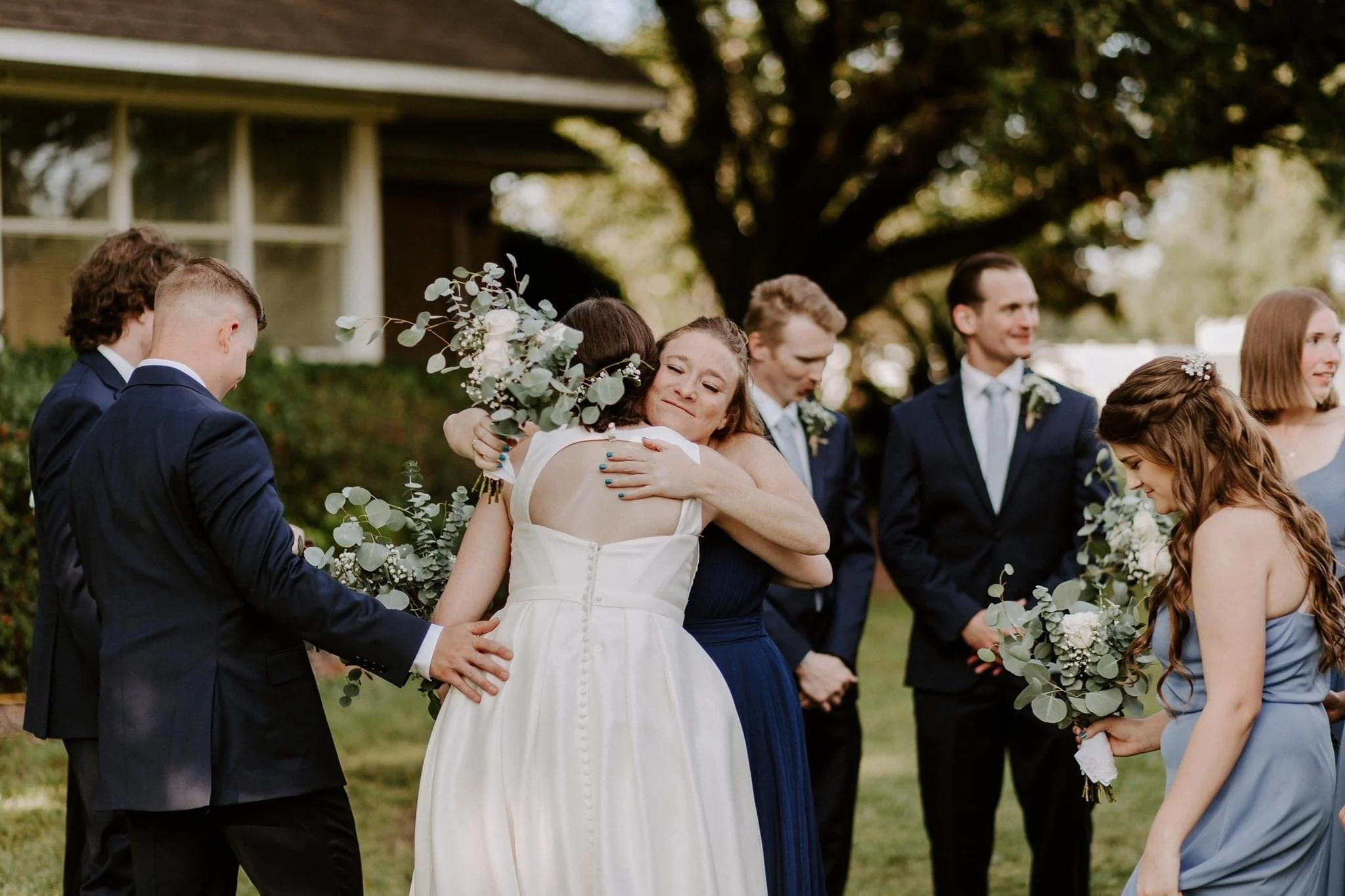 A woman in a white dress hugging another woman in a dark blue dress at an outdoor wedding, surrounded by bridesmaids and groomsmen holding bouquets.