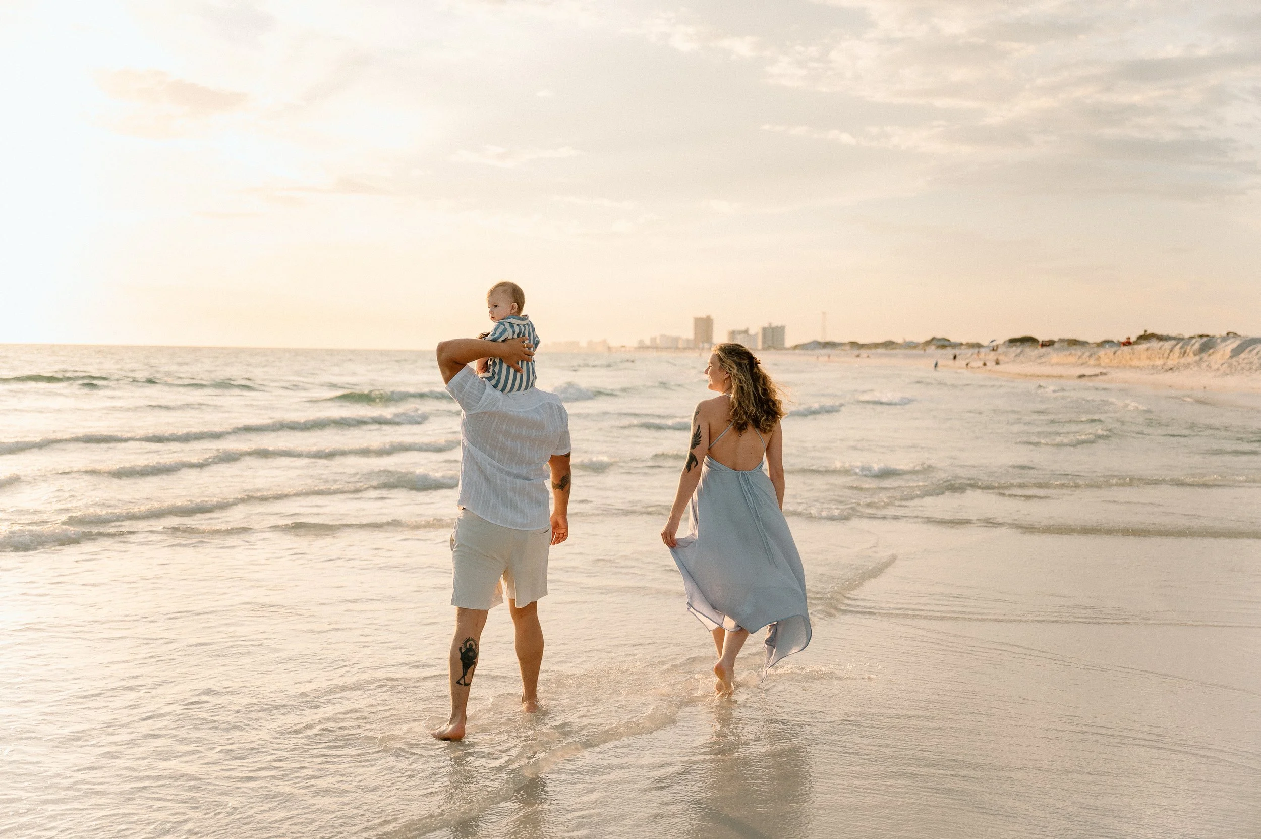 A family walking along the beach at sunset, with a man carrying a child on his shoulders and a woman walking beside them, all dressed in light, summery clothing.