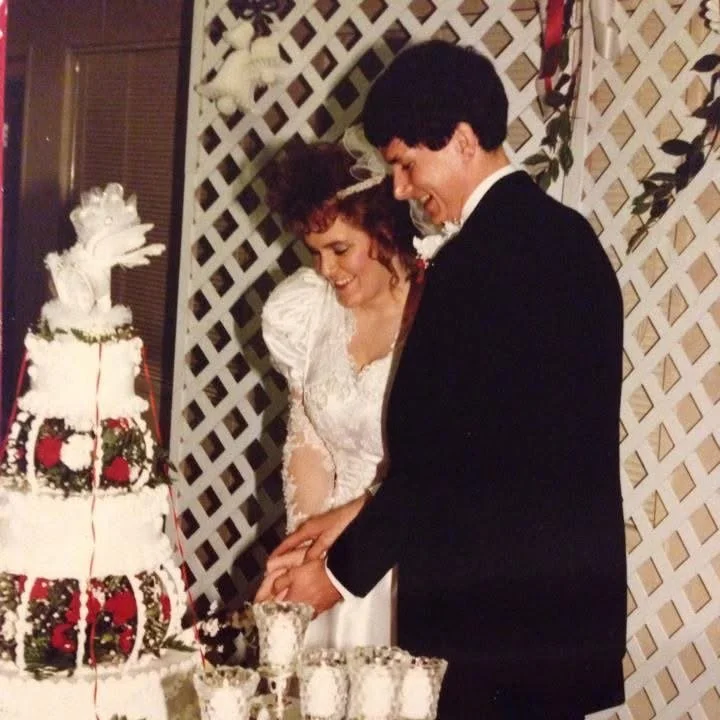 A bride and groom cutting their wedding cake, standing in front of a decorative lattice backdrop with white and red flowers.