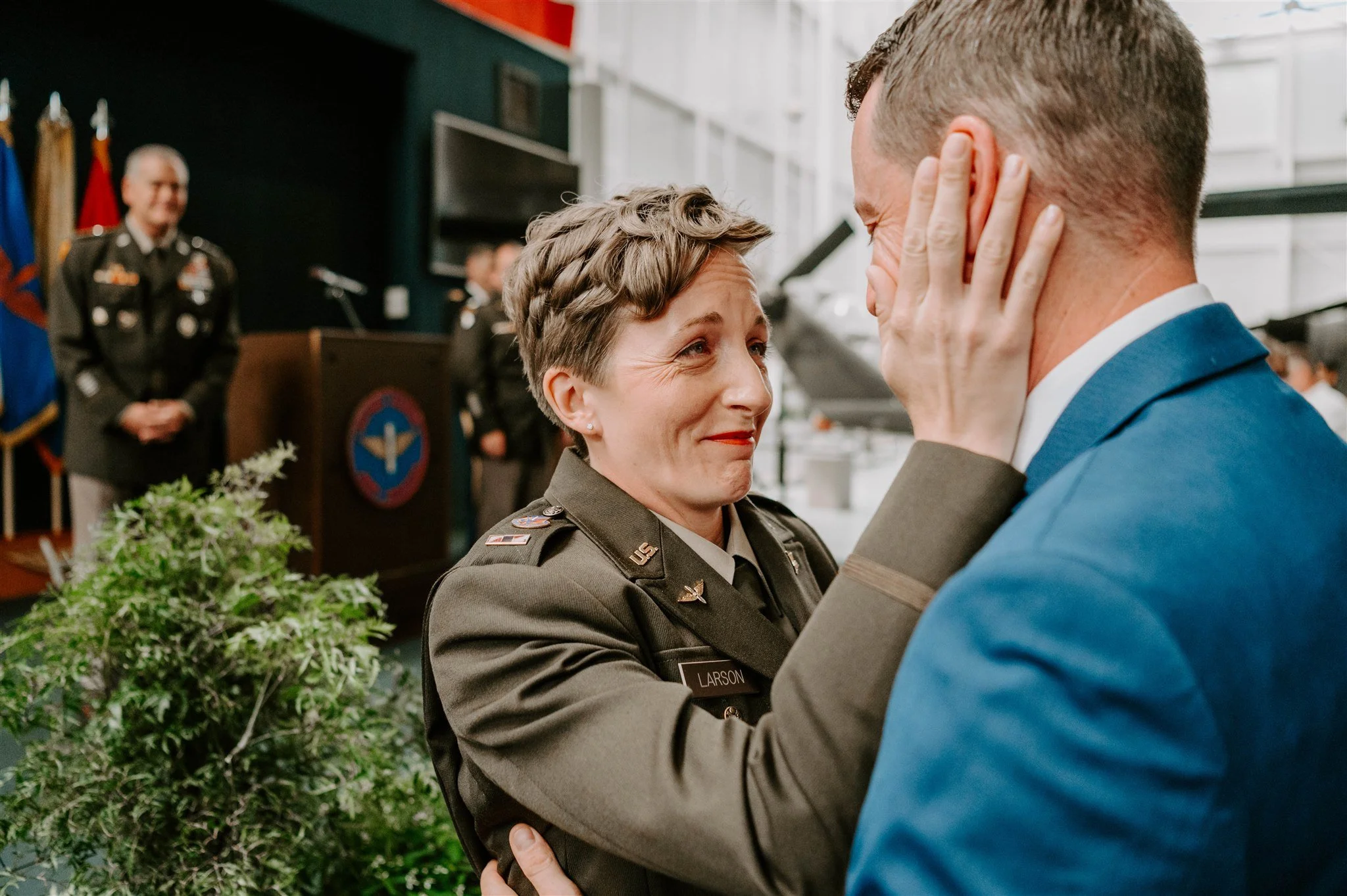A woman in military uniform is touching a man's face and smiling during a ceremony. Several people in military uniforms are in the background.