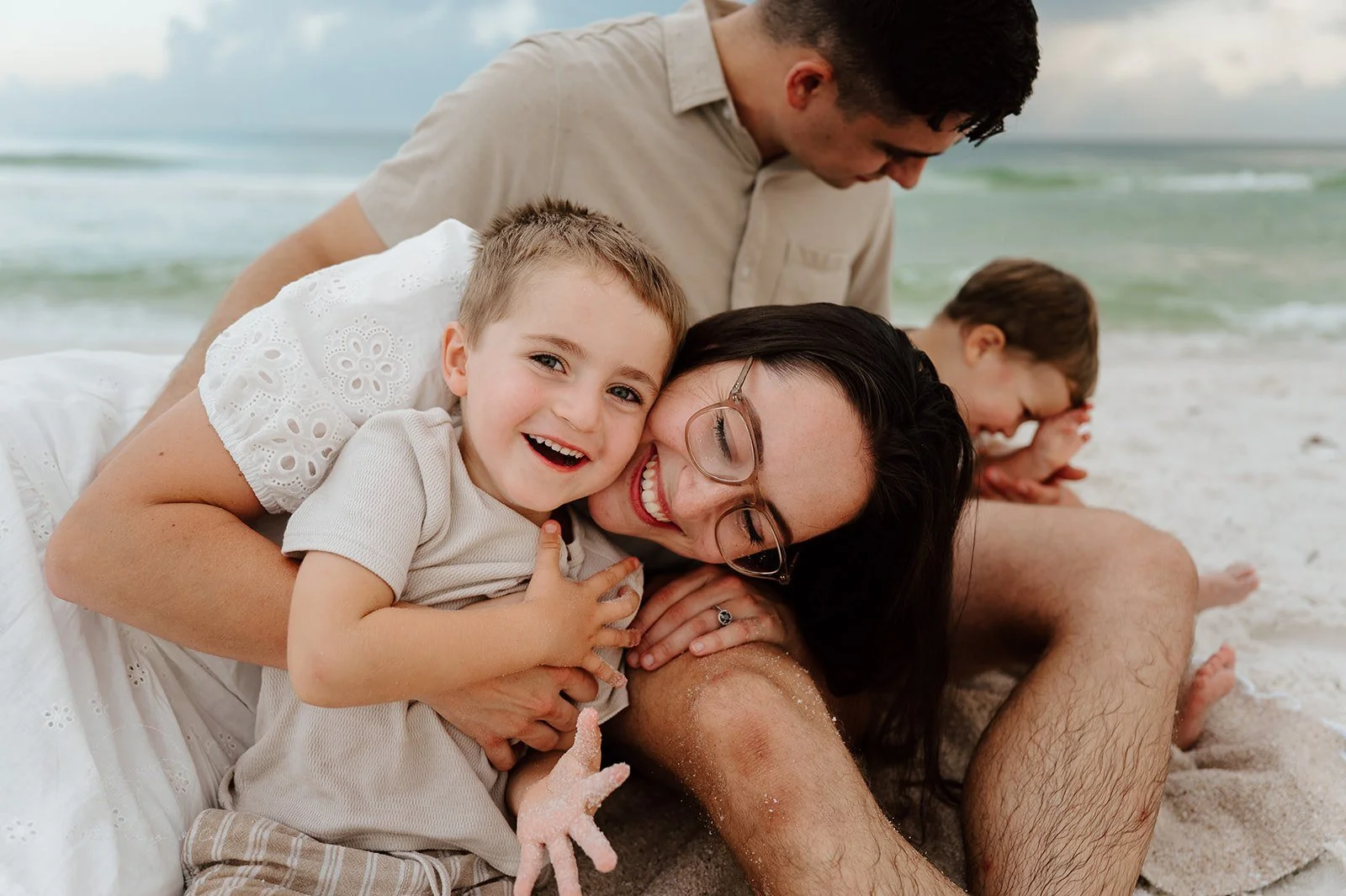 A family of four, including two children, sitting on the beach and enjoying a fun moment together. The woman and a young boy are smiling and embracing, with the woman holding a small pink toy. The man and girl are in the background, sitting close to 