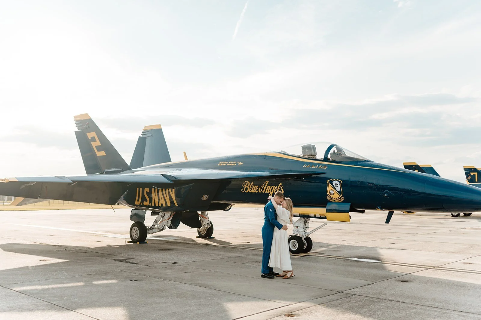 A couple in wedding attire stands in front of a U.S. Navy fighter jet on a tarmac, with the groom in a blue military uniform and the bride in a white dress, sharing a kiss.