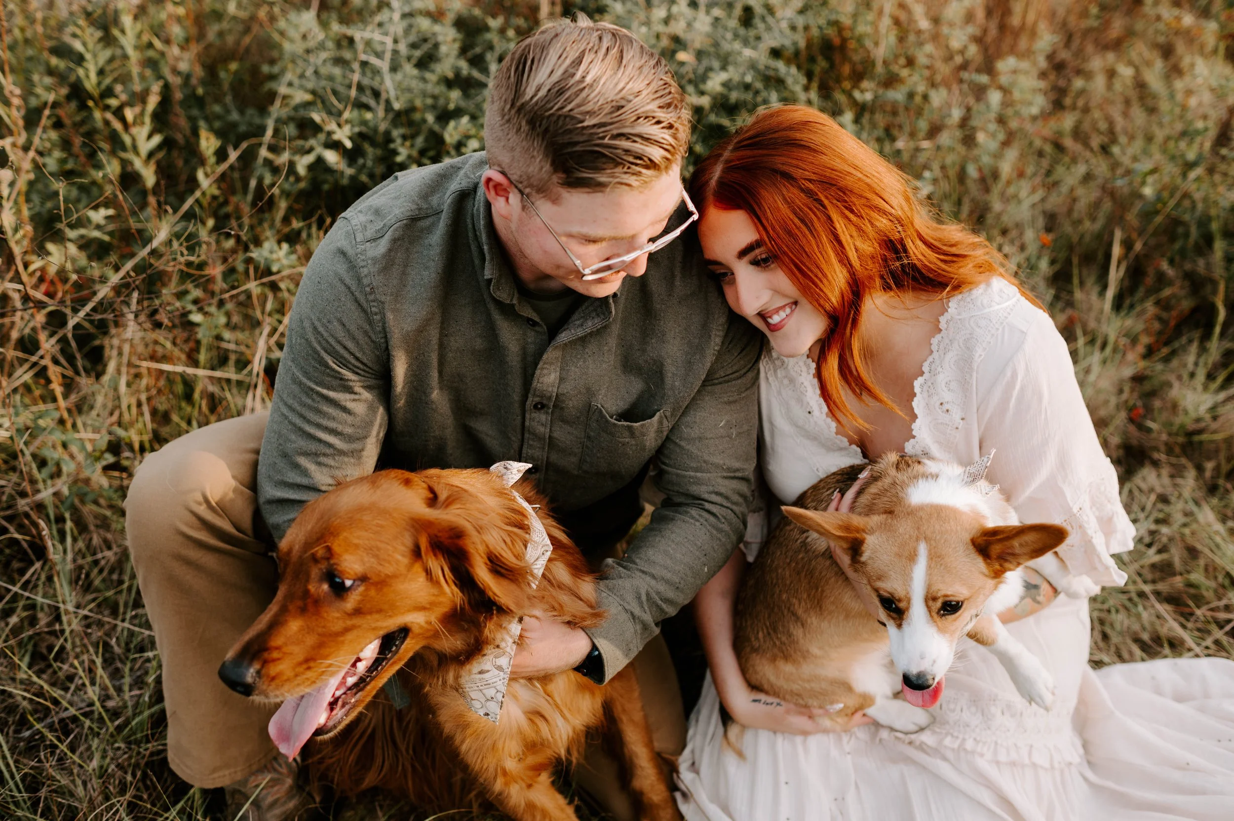 A couple sitting outdoors on grass, each holding a dog, smiling, surrounded by greenery and plants.
