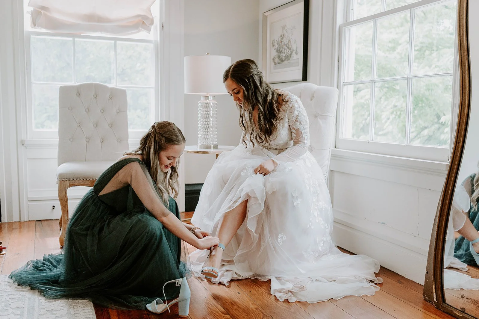 A bride in a white wedding dress sits on a white armchair while a bridesmaid in a dark green dress helps her put on a shoe in a bright room with large windows and wooden floors.