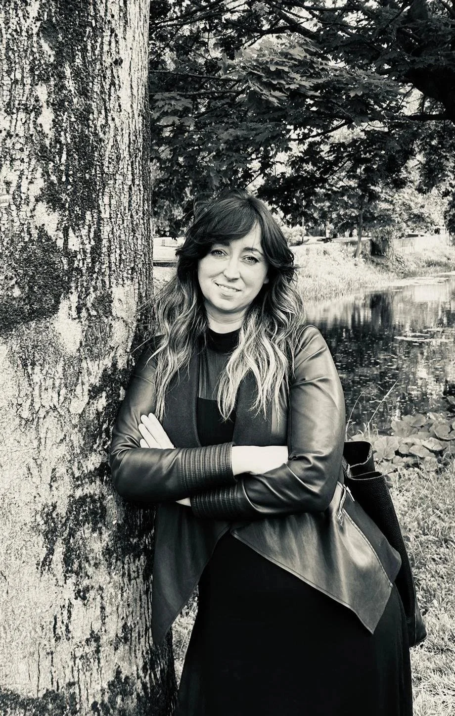 Interior Designer & Architect with dark hair, smiling, standing against a rocky wall outdoors. She is wearing a black top, a large beaded necklace, a white watch, and a crossbody bag.