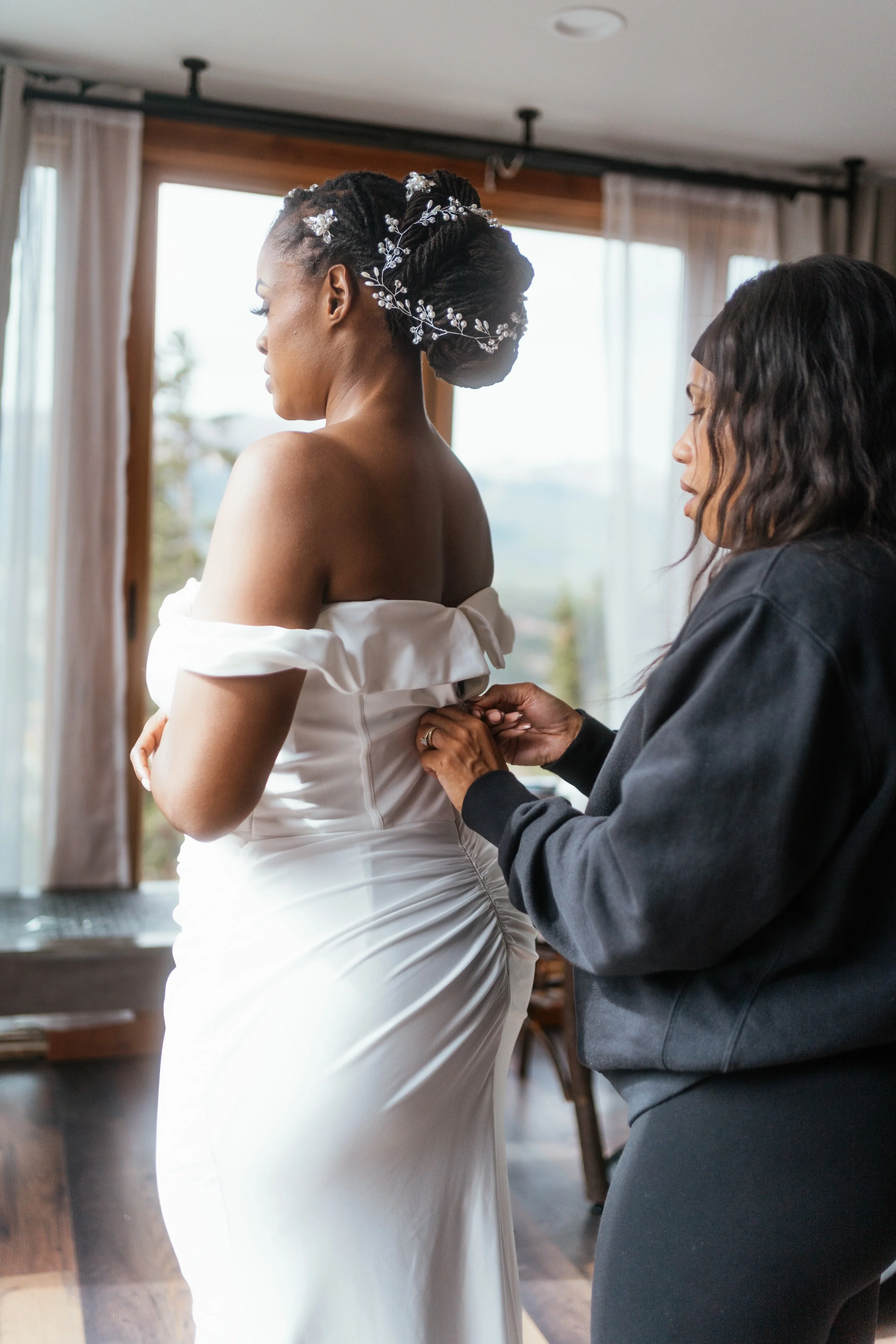 A bride gets ready at an airbnb while her hair and makeup artist snaps the last few buttons of her dress.