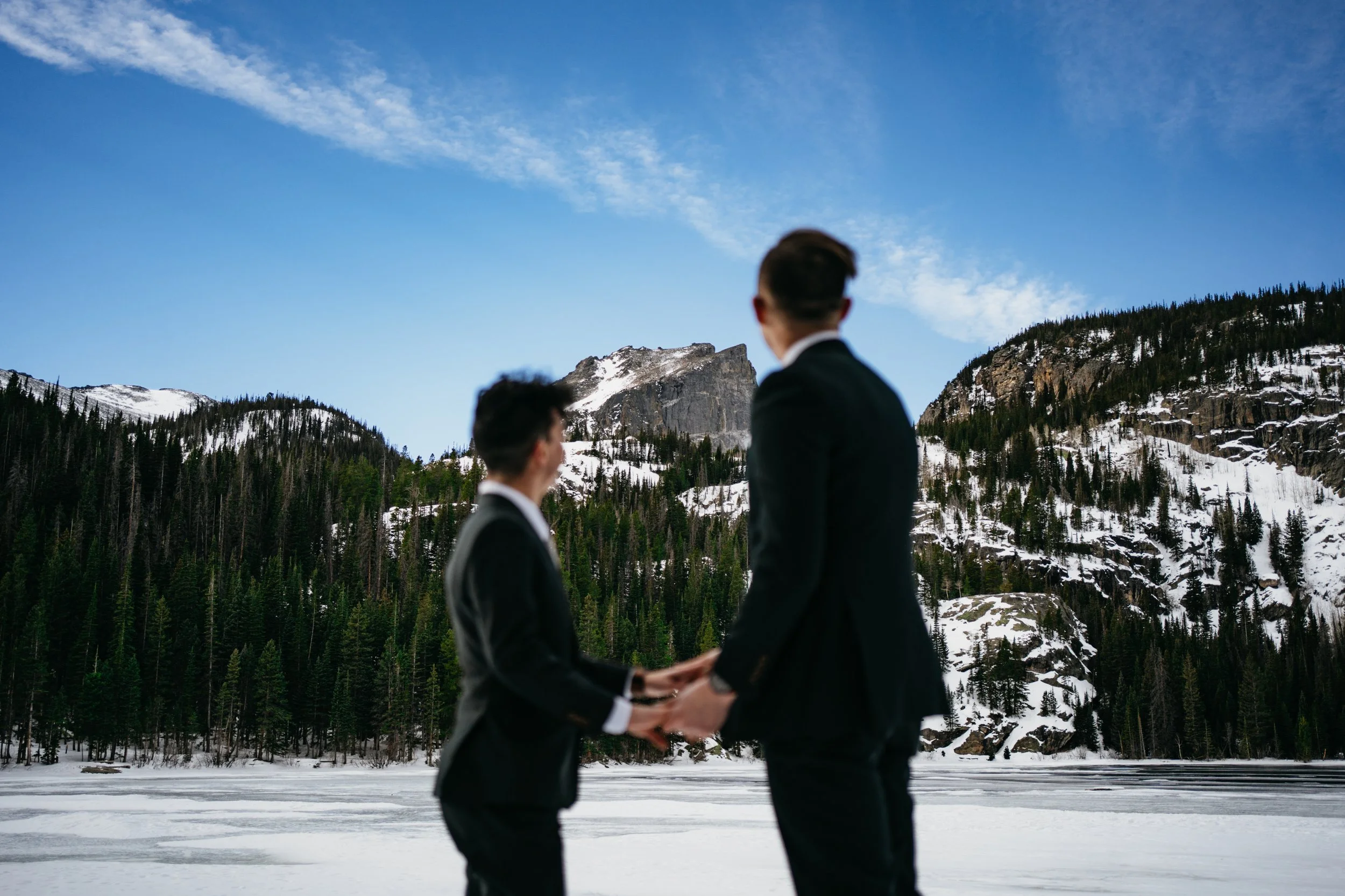 bear lake in rocky mountain national park iced over and covered in snow with two grooms holding hands in front of the lake.
