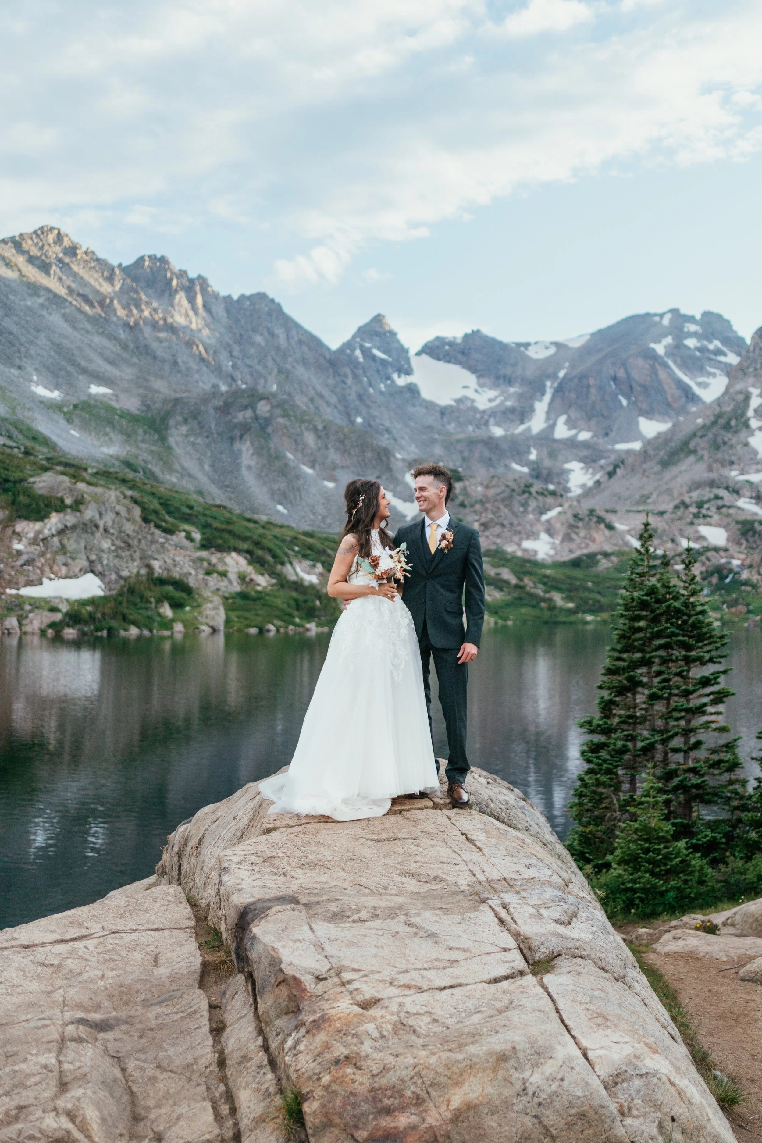 Couple standing on a rock beside an alpine lake beneath a lightly clouded sky