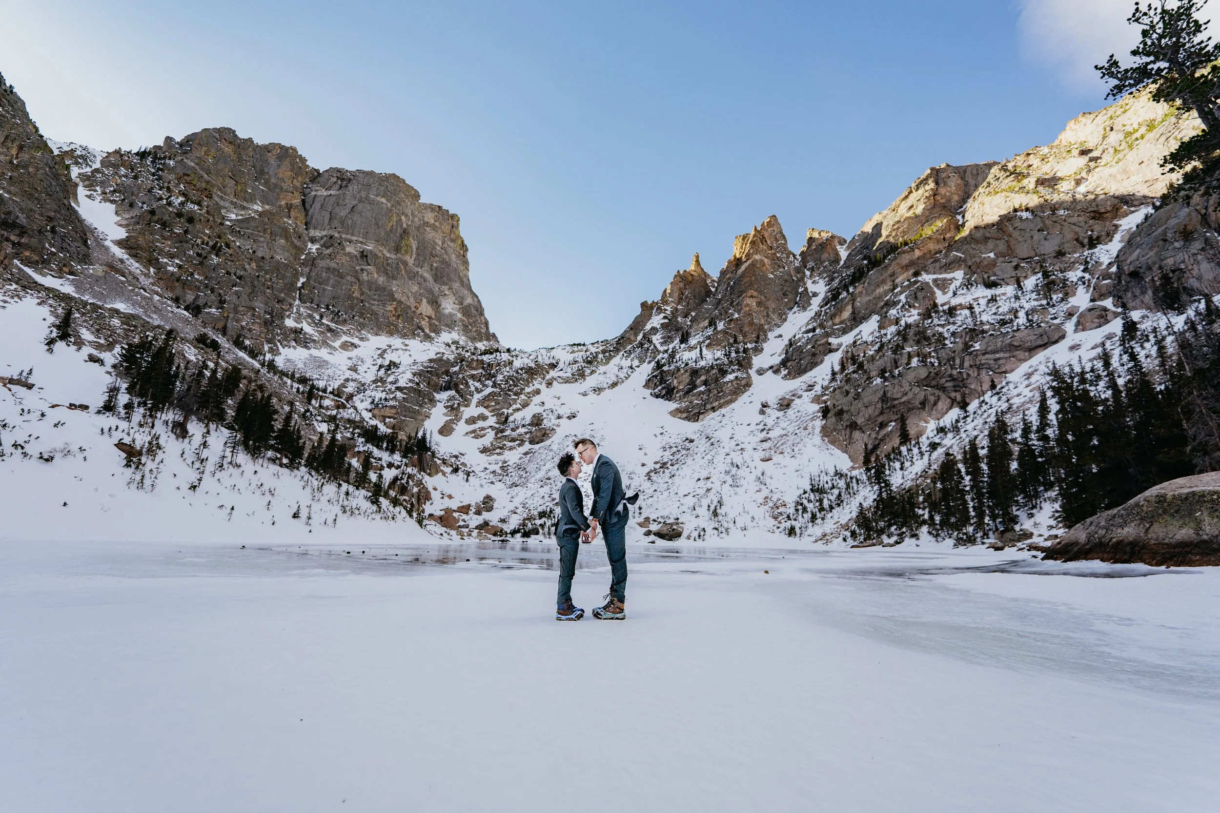 A groom and groom stand on an iced over lake about to kiss on their wedding day surrounded by the Colorado Rocky Mountains.